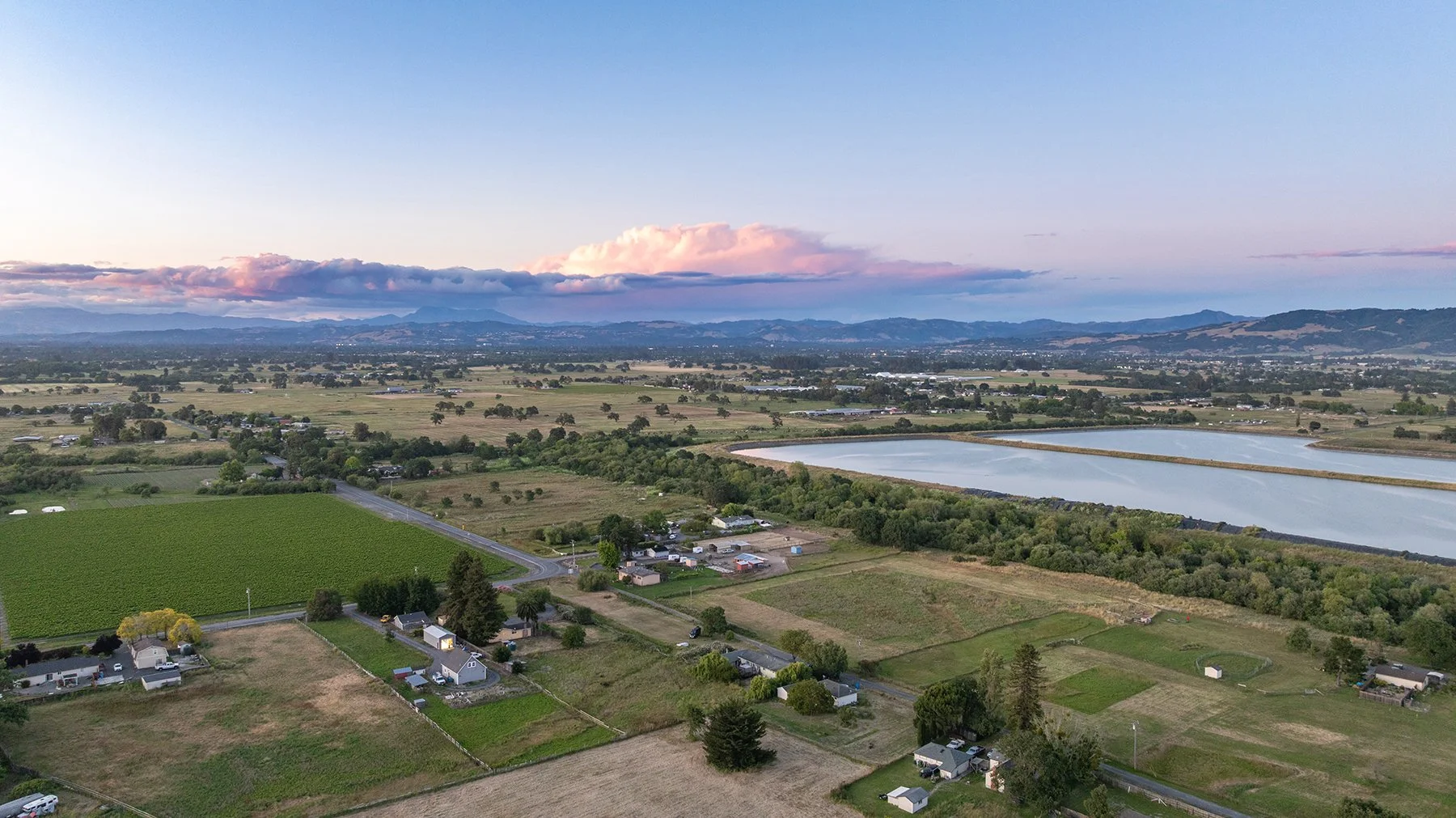 Aerial view of a rural landscape with fields, small houses, a large pond, and distant mountains under a partly cloudy sky.