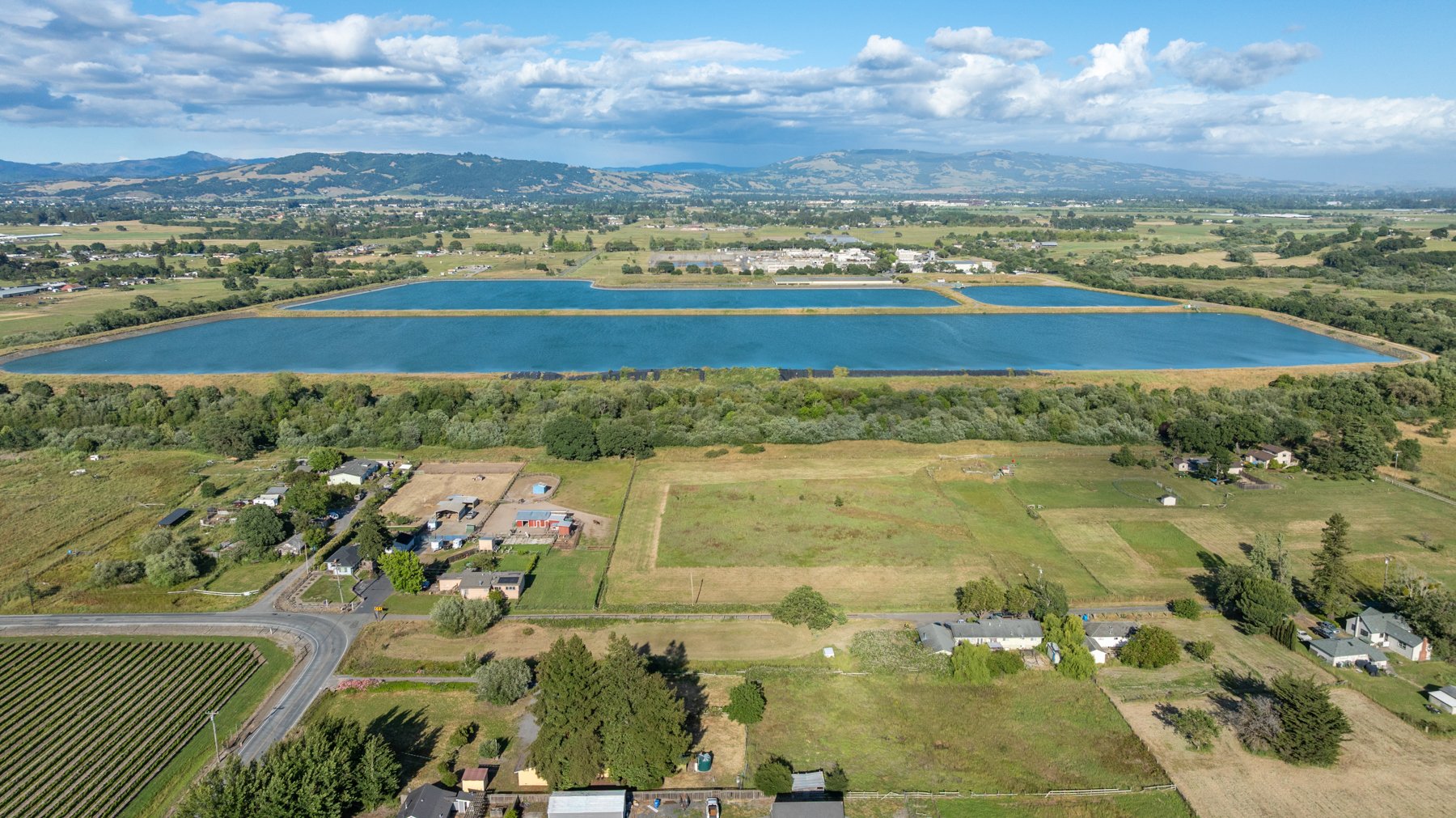 Aerial view of two large rectangular bodies of water, surrounded by farmland, houses, and trees with mountains in the distance under a partly cloudy sky.