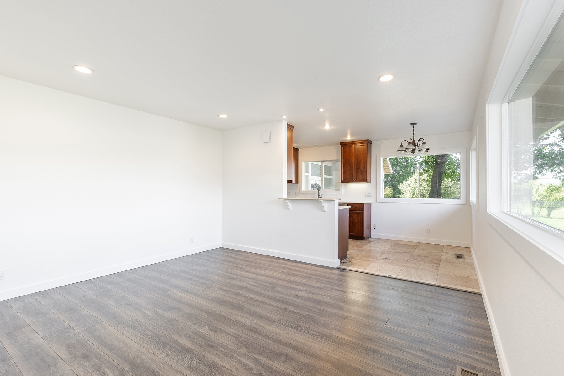 Empty living room with wood laminate flooring, white walls, large windows, and a view of greenery outside. The space is open to the kitchen with wooden cabinets and a tiled floor.