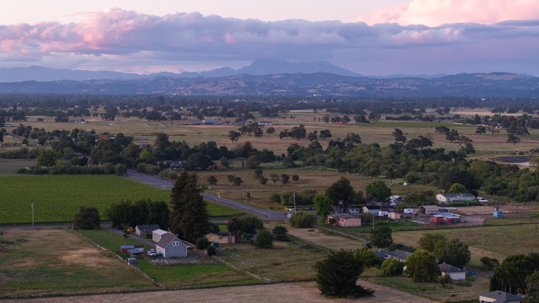 A rural landscape with green fields, scattered houses, and a winding road, with a mountain range in the distance under a partly cloudy sky at sunset.
