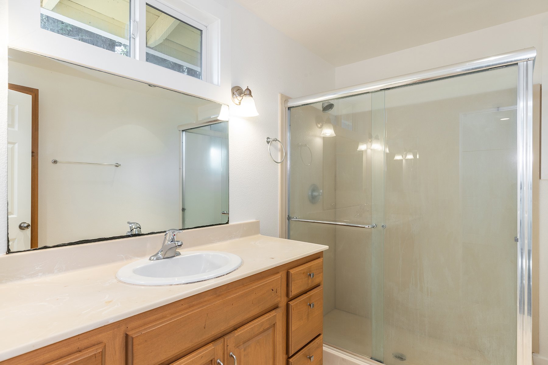 Bathroom with wooden vanity, oval sink, large mirror, glass shower door, and small window above mirror.