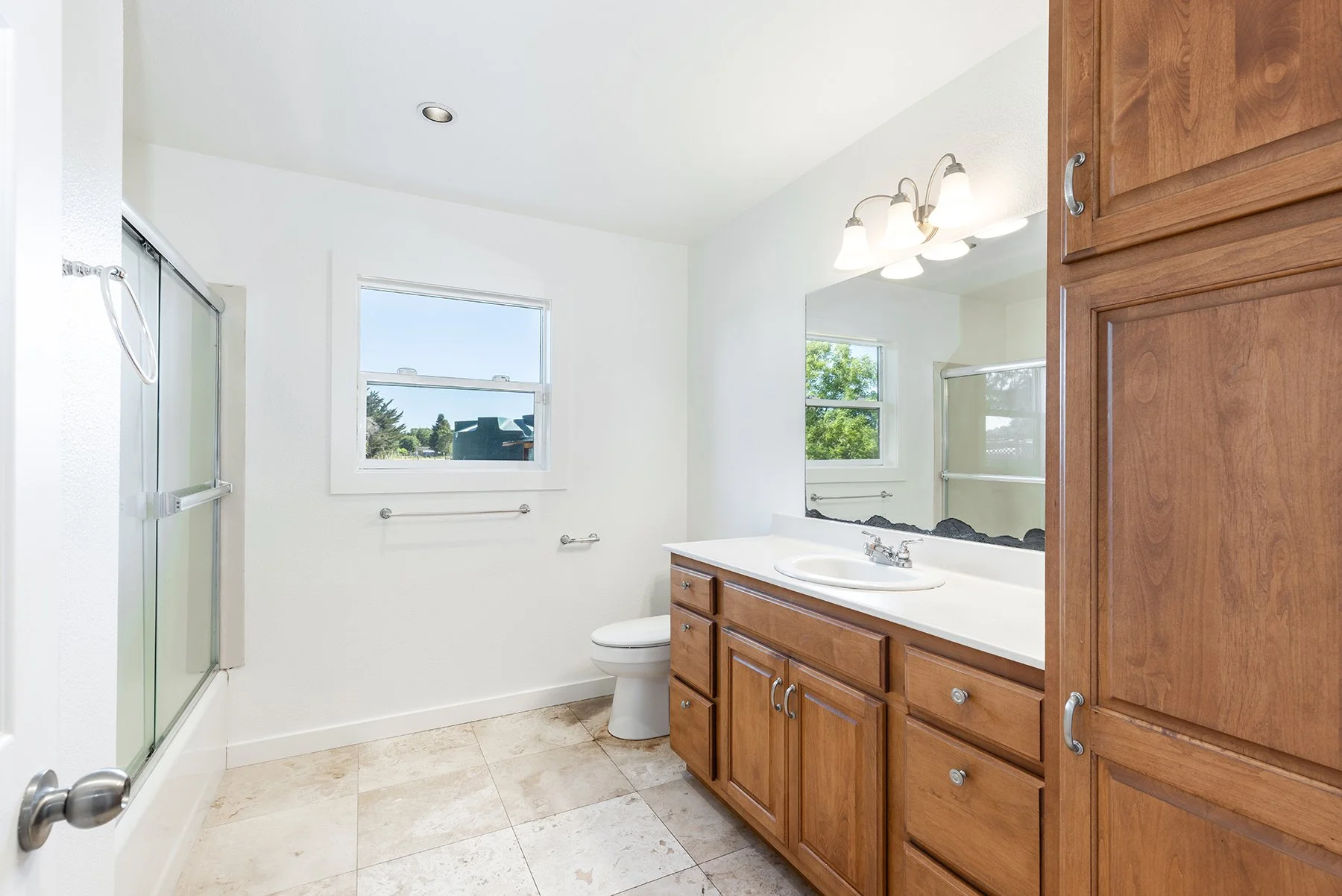 Bathroom with white walls, beige tiled floor, wooden cabinetry, a white sink with a chrome faucet, a large wall mirror, a window showing blue sky and trees, a toilet, a shower with a glass door, and ceiling lights.