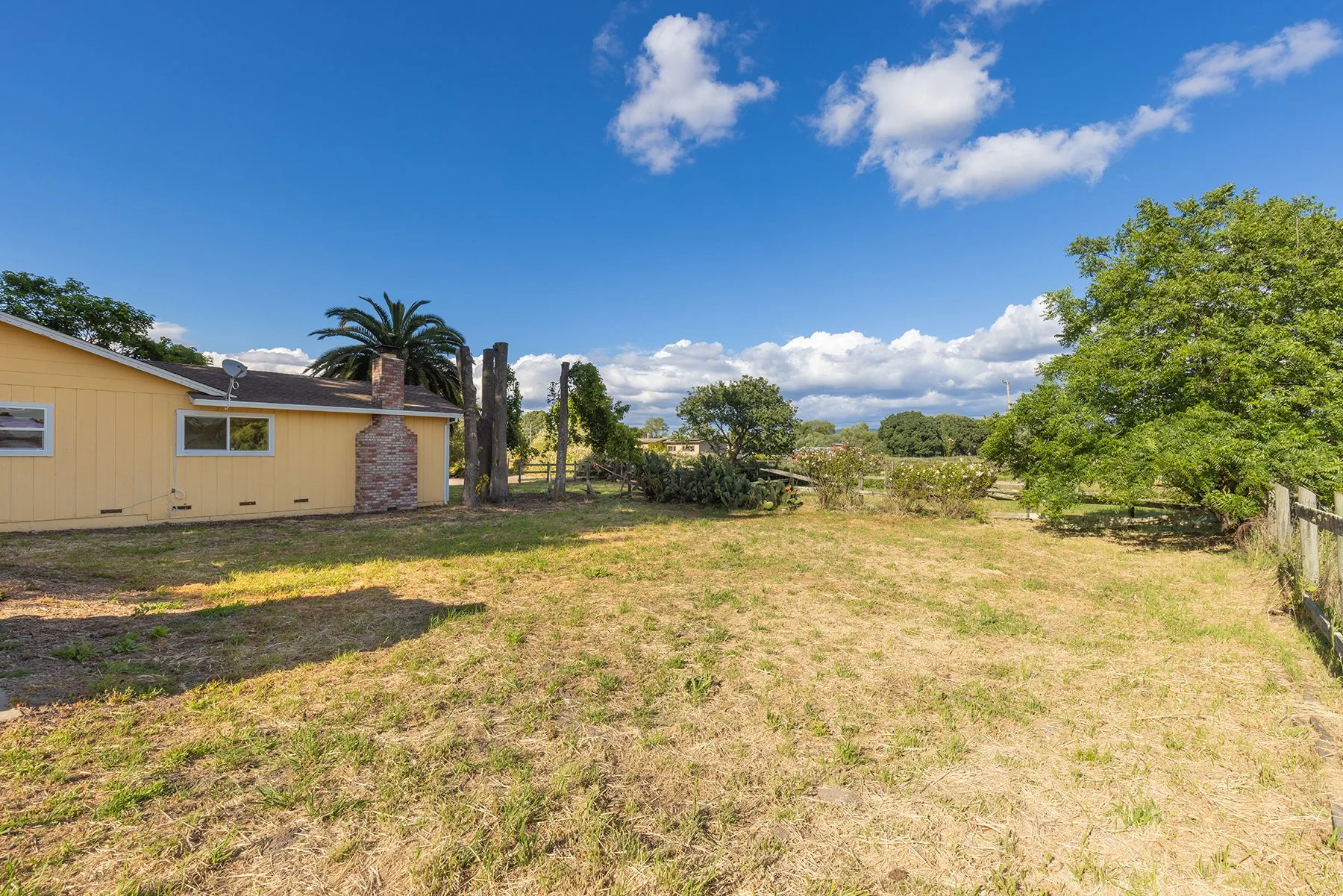 Residential backyard with a yellow house, trees, and a clear blue sky with scattered clouds.