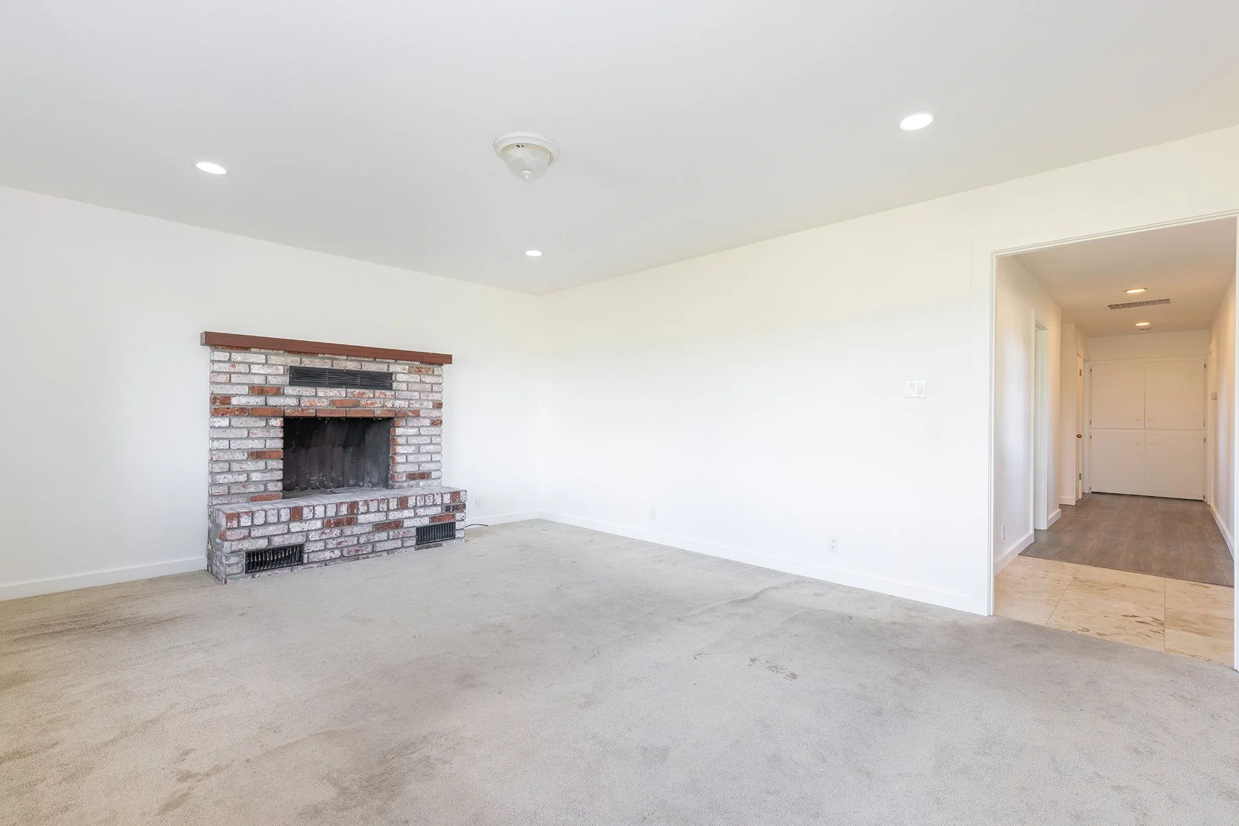 Empty living room with brick fireplace, white walls, and a hallway leading to a white door.