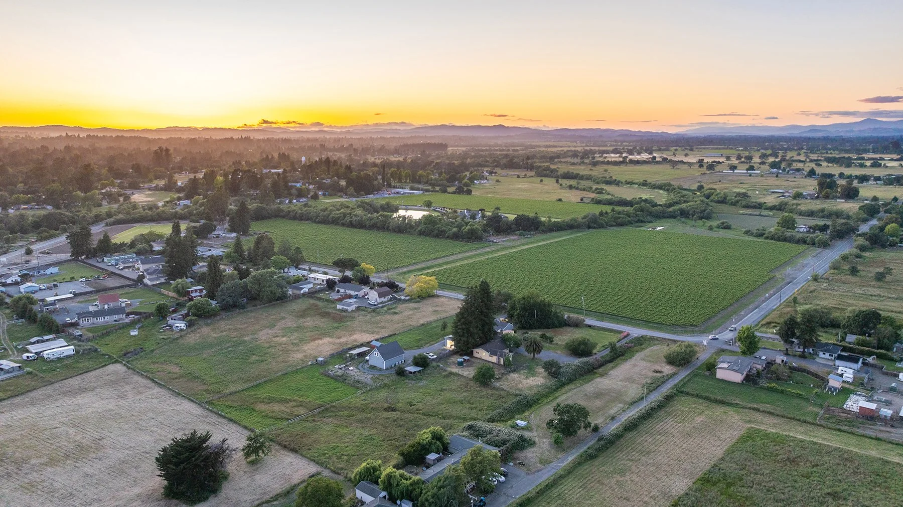 Aerial view of a rural landscape at sunset showing farmland, small houses, roads, and a forest in the distance.