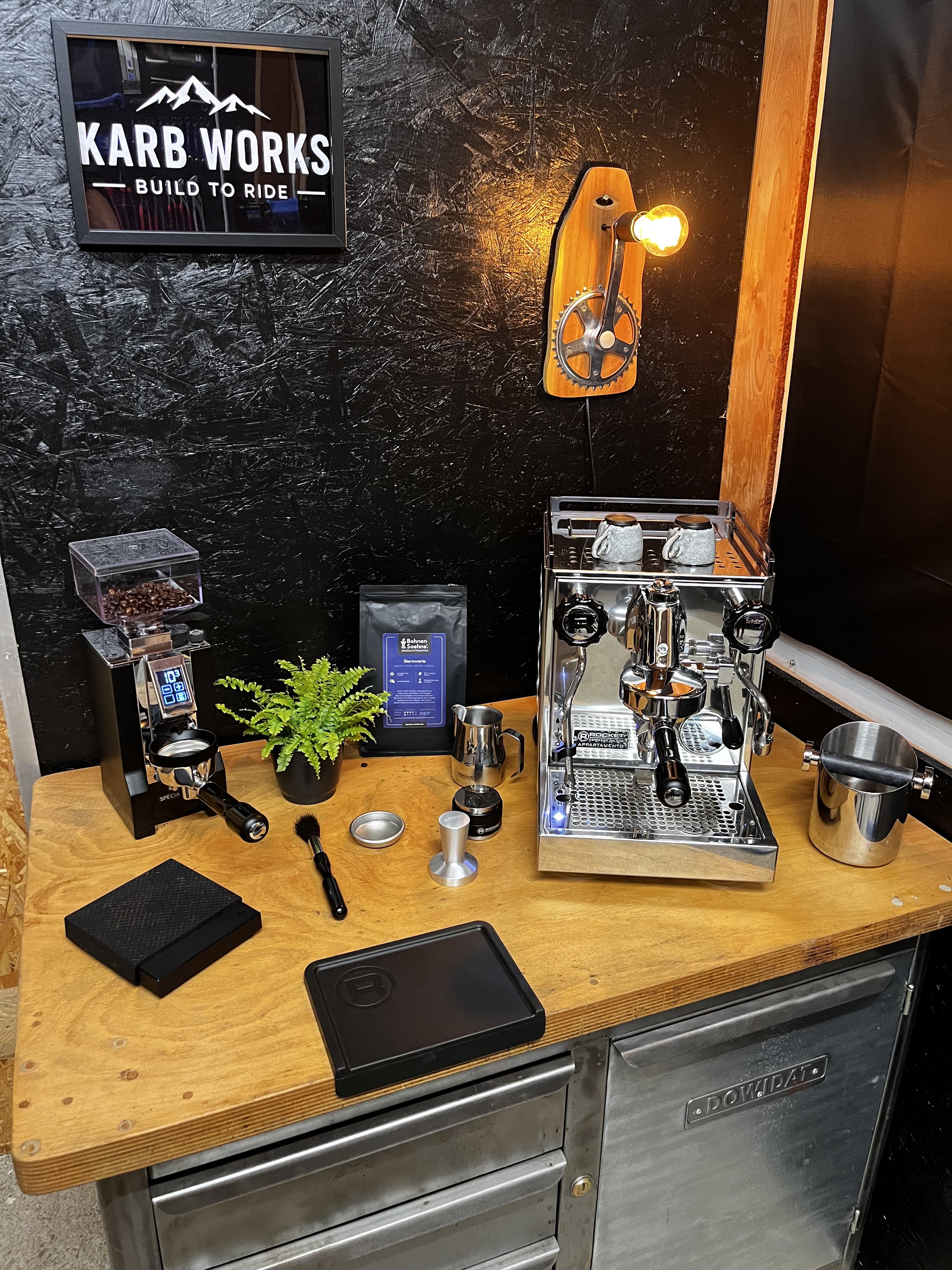 Ein Kaffeezubereitungsbereich mit Espressomaschine, Kaffeetassen, Kaffeebohnen, einem Messer, einem kleinen Pflanze, einer Waage, einem schwarzen Notizbuch, einem Stift und einer Wärmetasche auf einer Holztisch. Im Hintergrund eine schwarze Wand und ein Wandbild mit dem Text „KARRB WORKS BUILD TO RIDE“.