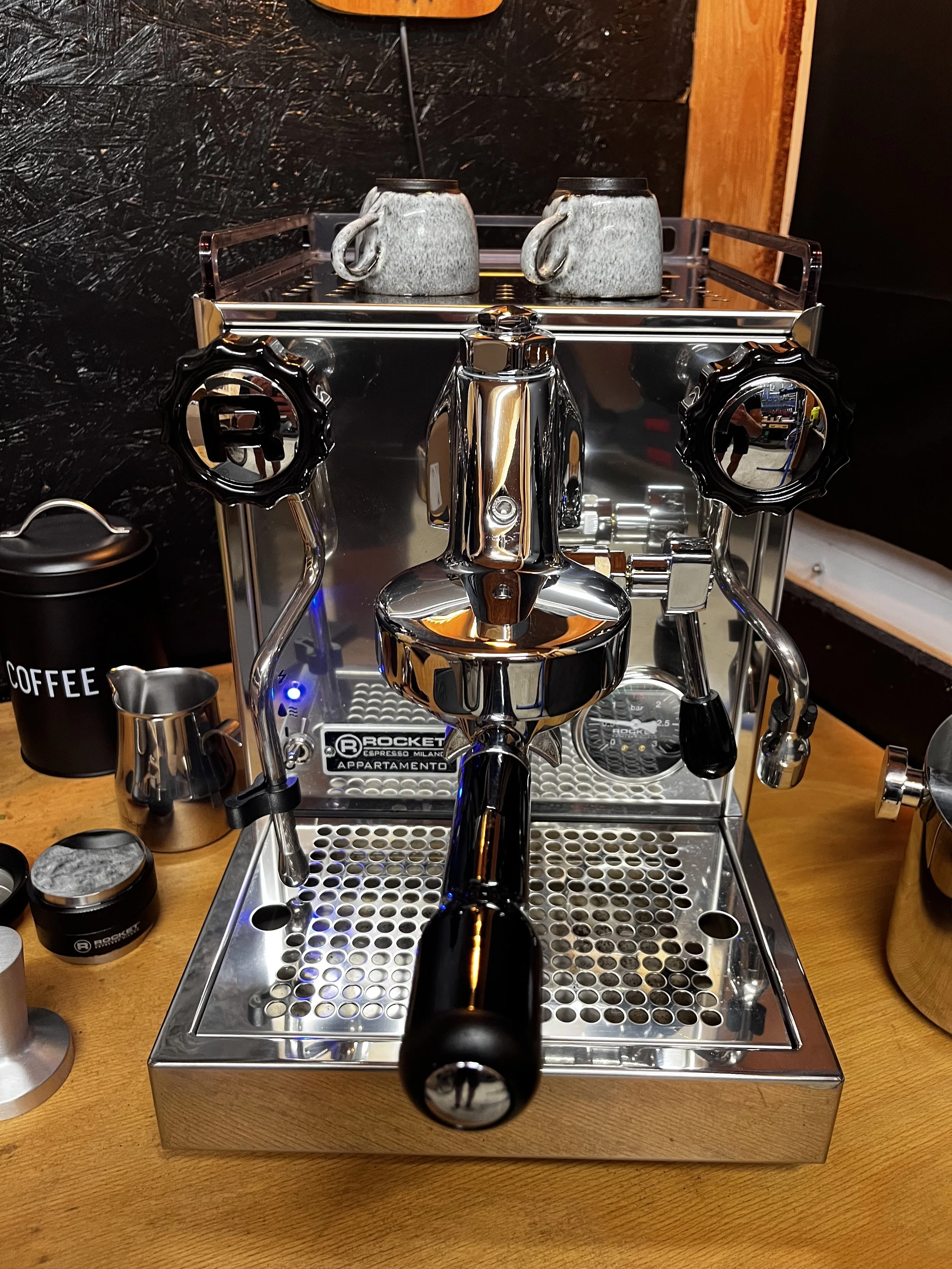 Ein Espressomaschine auf einem Holztablett mit zwei grauen Kaffeetassen auf der Oberseite, eines mit einem Strohhalm. Es gibt auch einen schwarzen Behälter mit weißen Buchstaben 'COFFEE' und einige kleine Behälter auf dem Tisch.