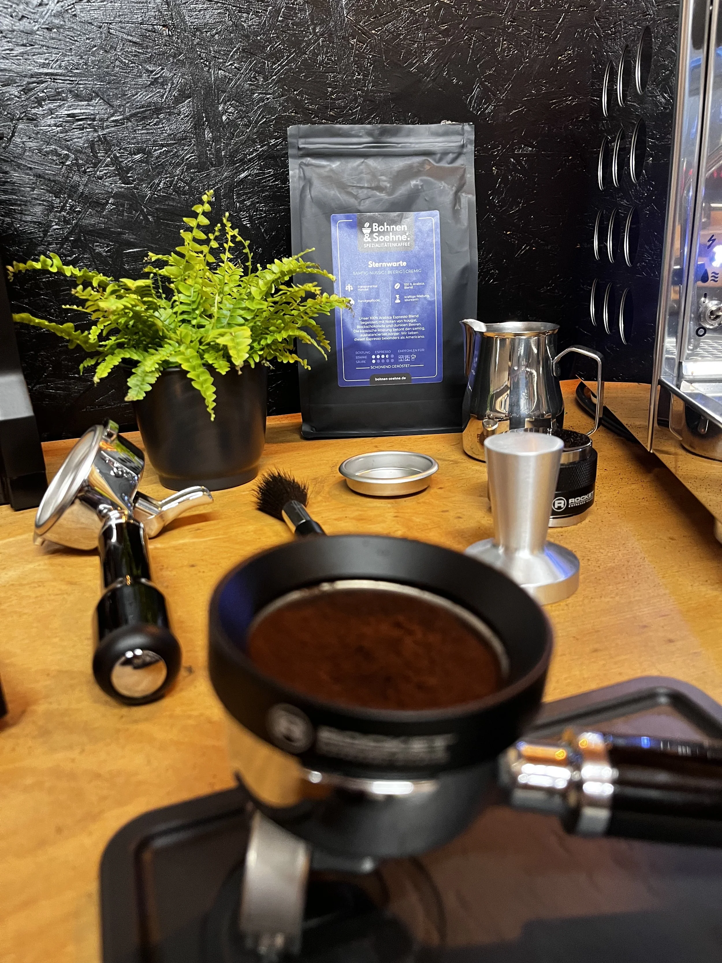 Ein Kaffeebereiter-Setup mit einer Kaffeepackung, einer Pflanze, einem Kaffeekännchen, einem Kaffeemühle, einer Tasse, einem Sieb und einem Kaffee-Filter auf einem Holztisch, im Hintergrund eine schwarze Wand.