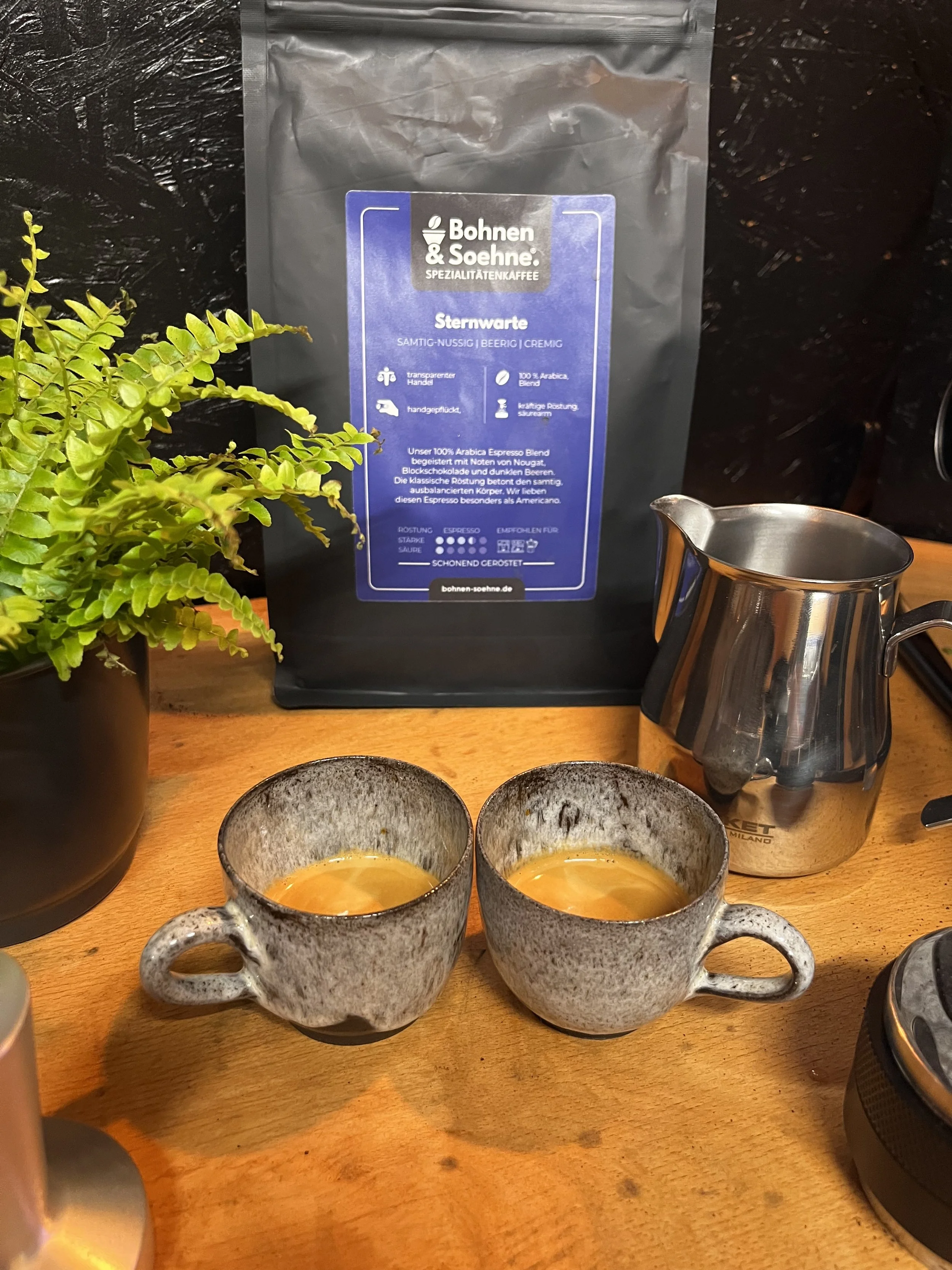 Zwei gefüllte graue Keramik-Tassen mit Kaffee, auf einem Holztisch, neben einem Zierpflanzenkübel, eine Kaffeemaschine, eine Tüte Gemahlenkaffee.