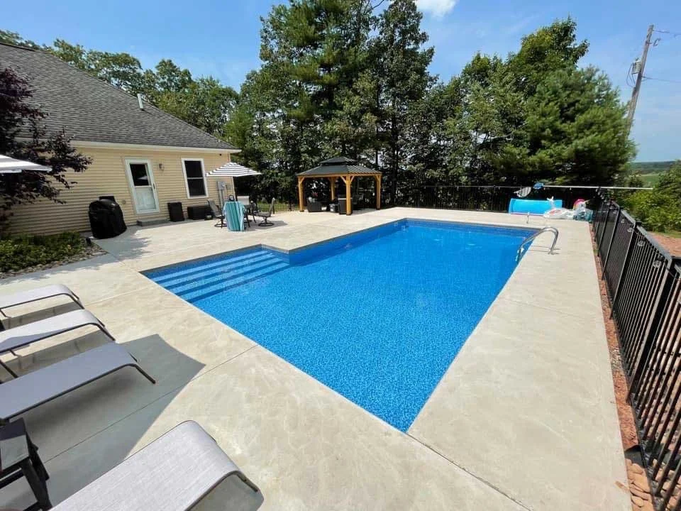Rectangular in-ground pool with wide entry steps, concrete pool deck, and backyard gazebo.