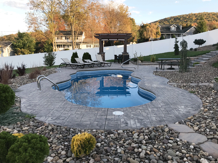 Custom freeform in-ground swimming pool with stamped concrete patio, landscaped rock beds, lounge seating, and pergola in a residential backyard.