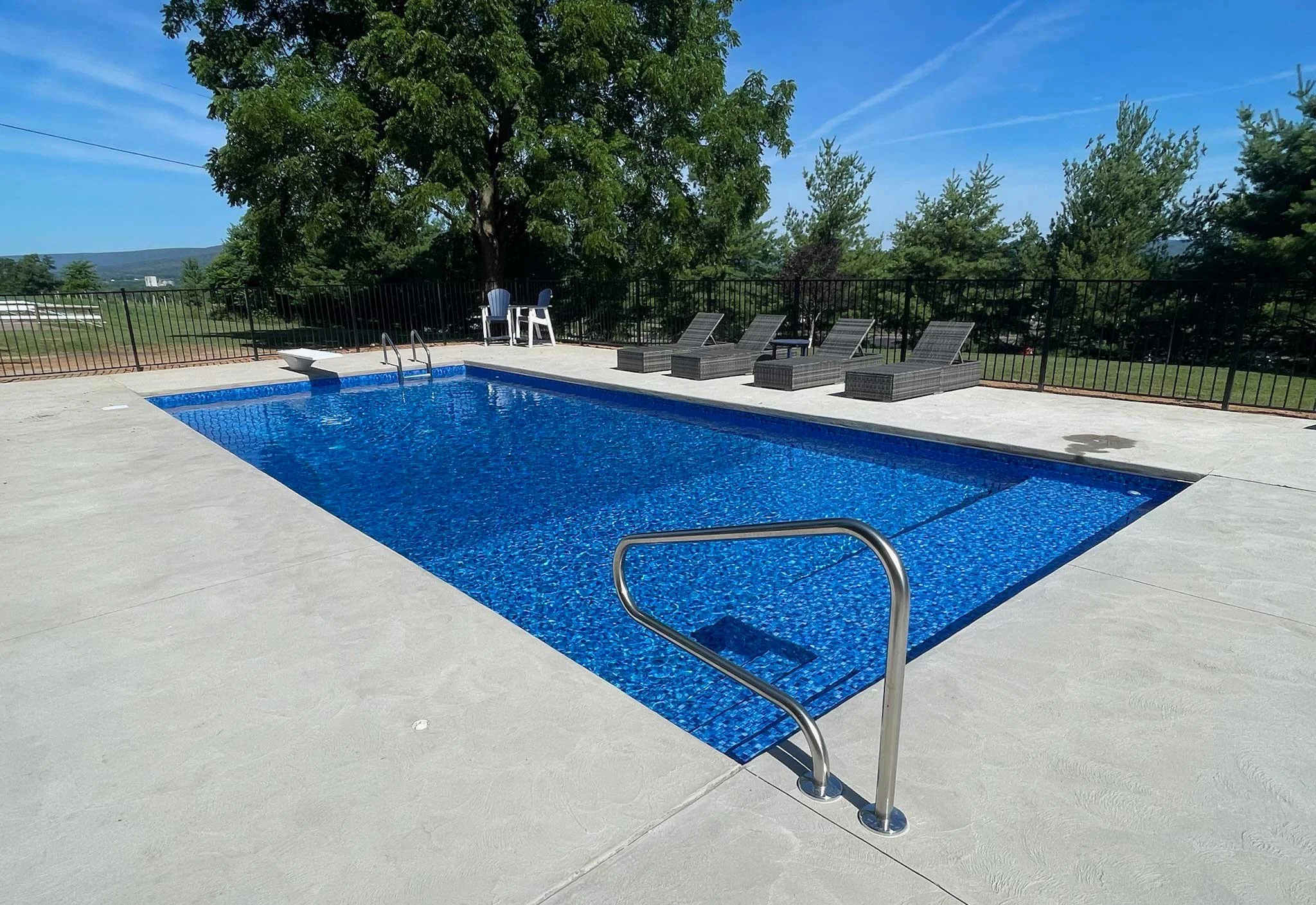 Rectangular in-ground pool with concrete decking, lounge seating, and black aluminum fencing.