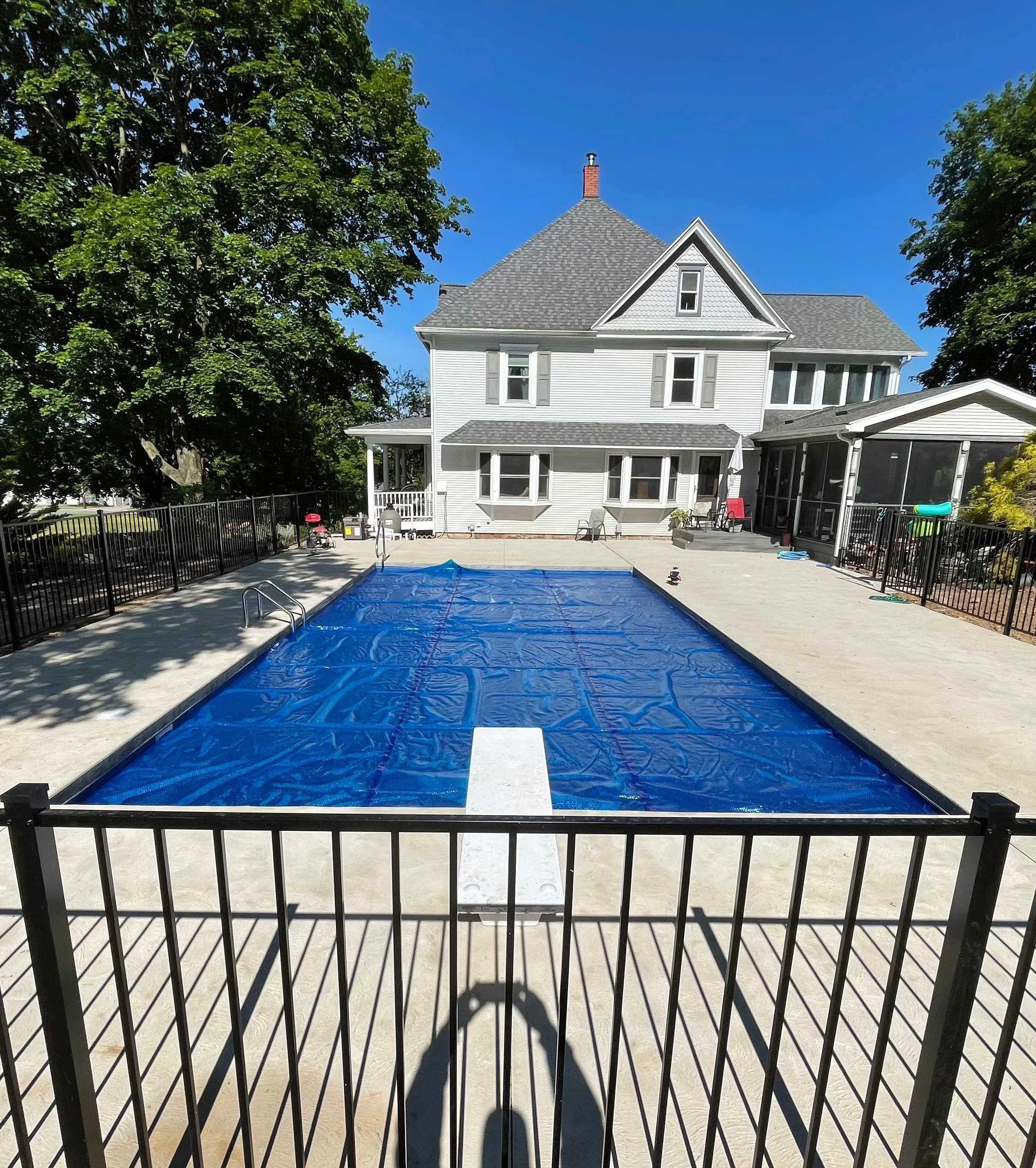 Rectangular in-ground swimming pool with safety cover, concrete decking, black aluminum fencing, and a two-story home in the background.
