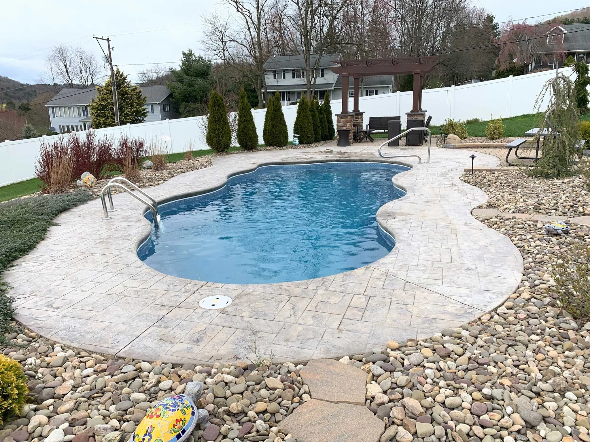 Freeform backyard swimming pool with stamped concrete surround, pergola, and landscaped rock beds