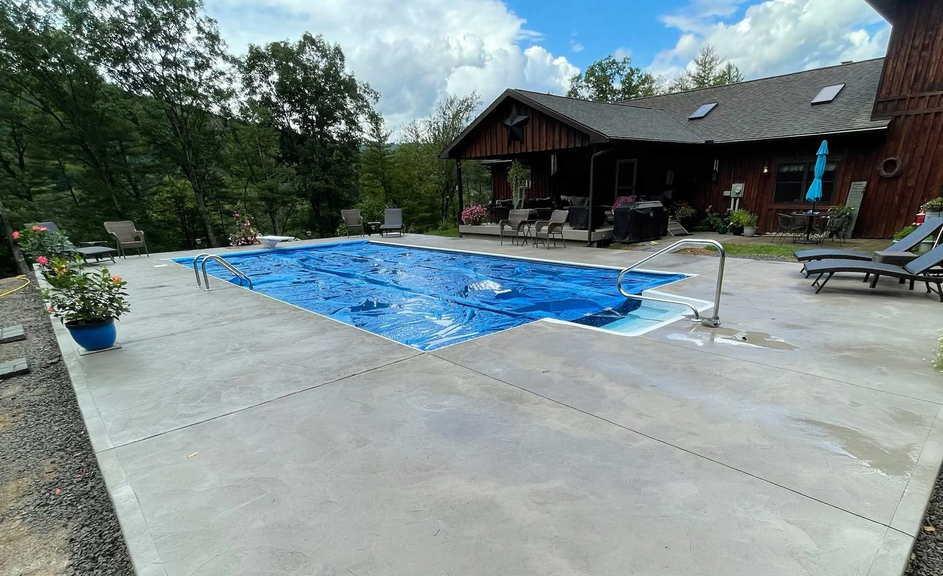 Rectangular in-ground swimming pool with cover beside a rustic home and wooded landscape