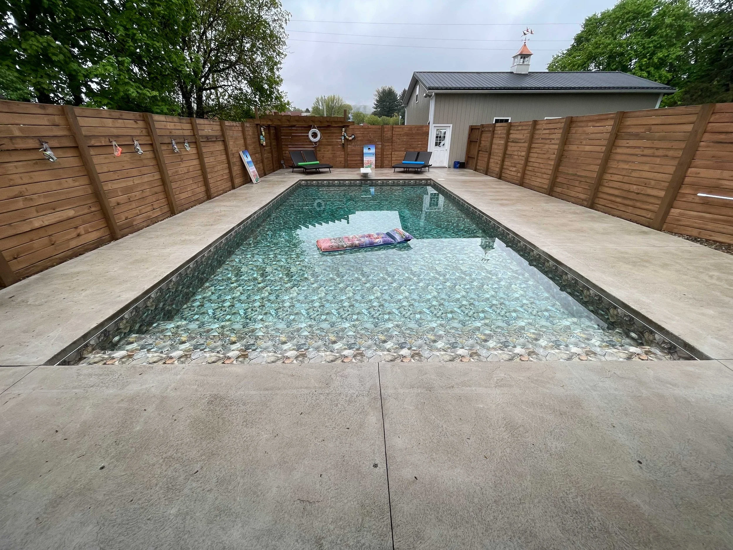 Rectangular backyard swimming pool with concrete decking, privacy fencing, and lounge seating.