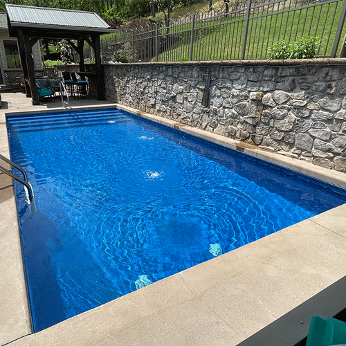 In-ground swimming pool surrounded by a stone retaining wall and landscaped backyard.