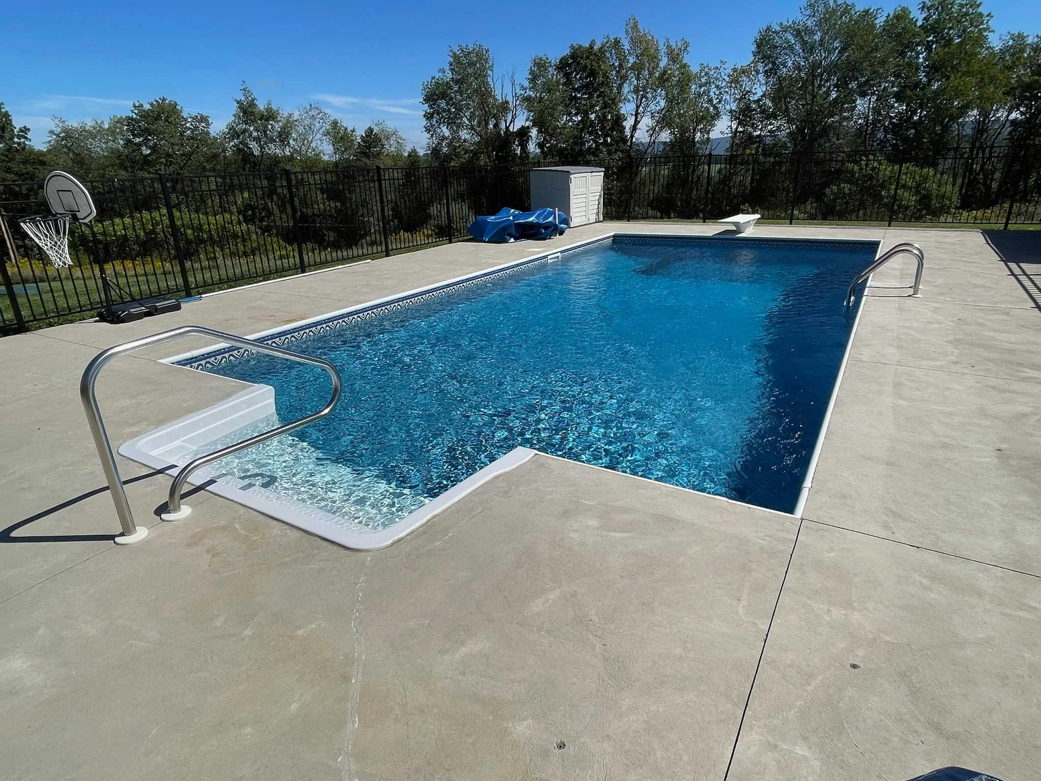 Rectangular in-ground swimming pool with diving board, basketball hoop, and expansive concrete deck