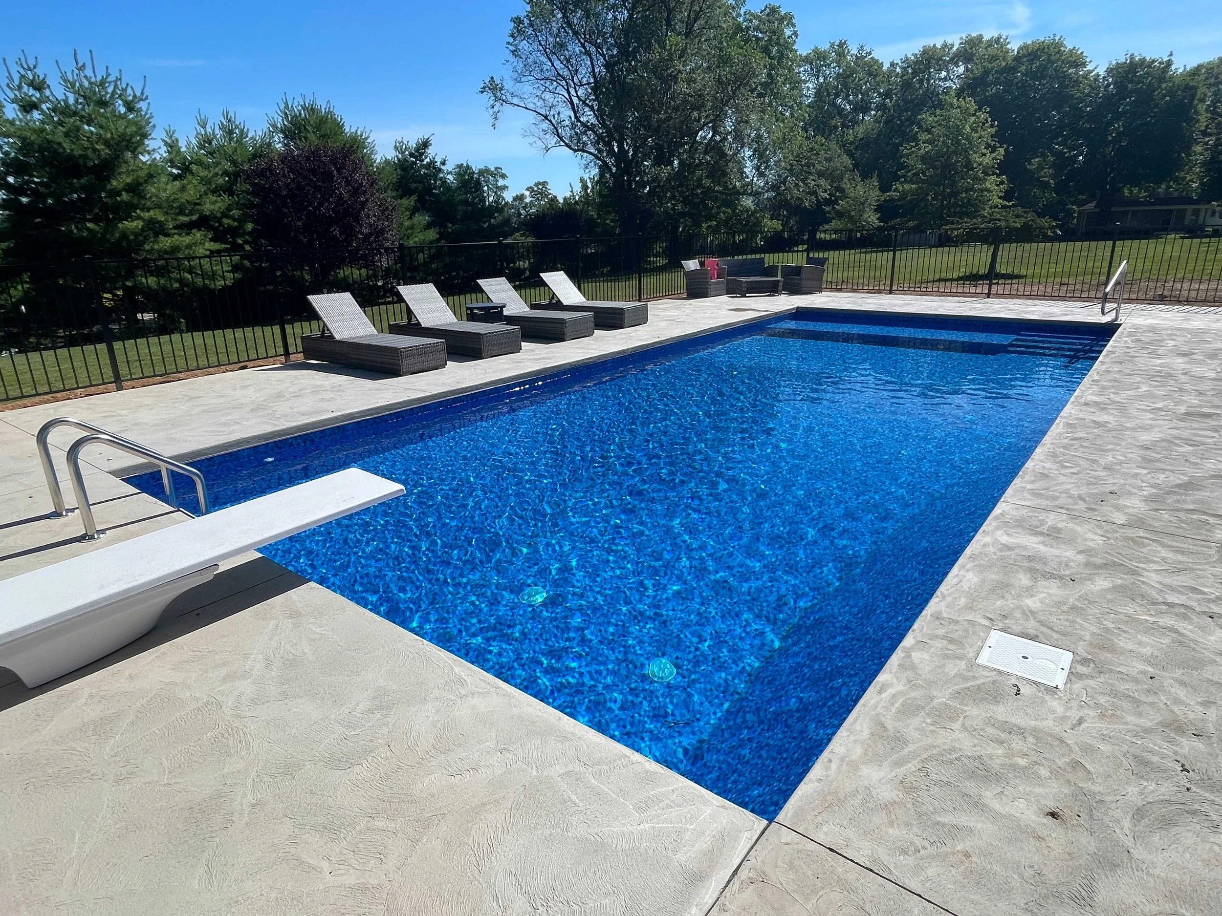 Rectangular in-ground swimming pool with concrete decking and backyard lounge area.