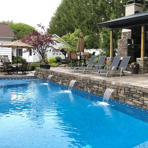 Luxury backyard pool featuring stone water features, outdoor fireplace, and spacious patio seating.
