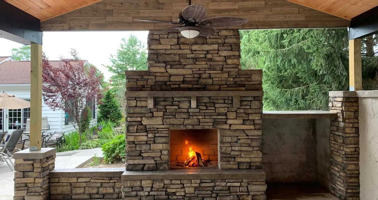 Covered outdoor living space featuring a custom stone fireplace overlooking a backyard pool area