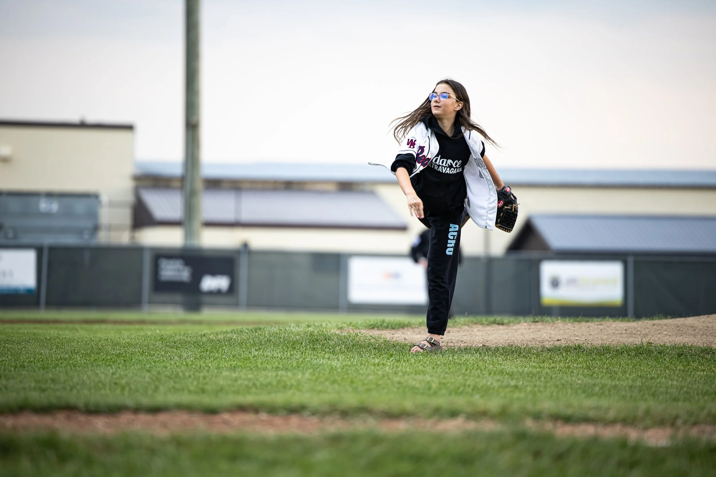 A young woman is standing on a baseball field, wearing a black t-shirt, black pants, and sunglasses. She is holding a baseball glove in her left hand and appears to be looking to her left.