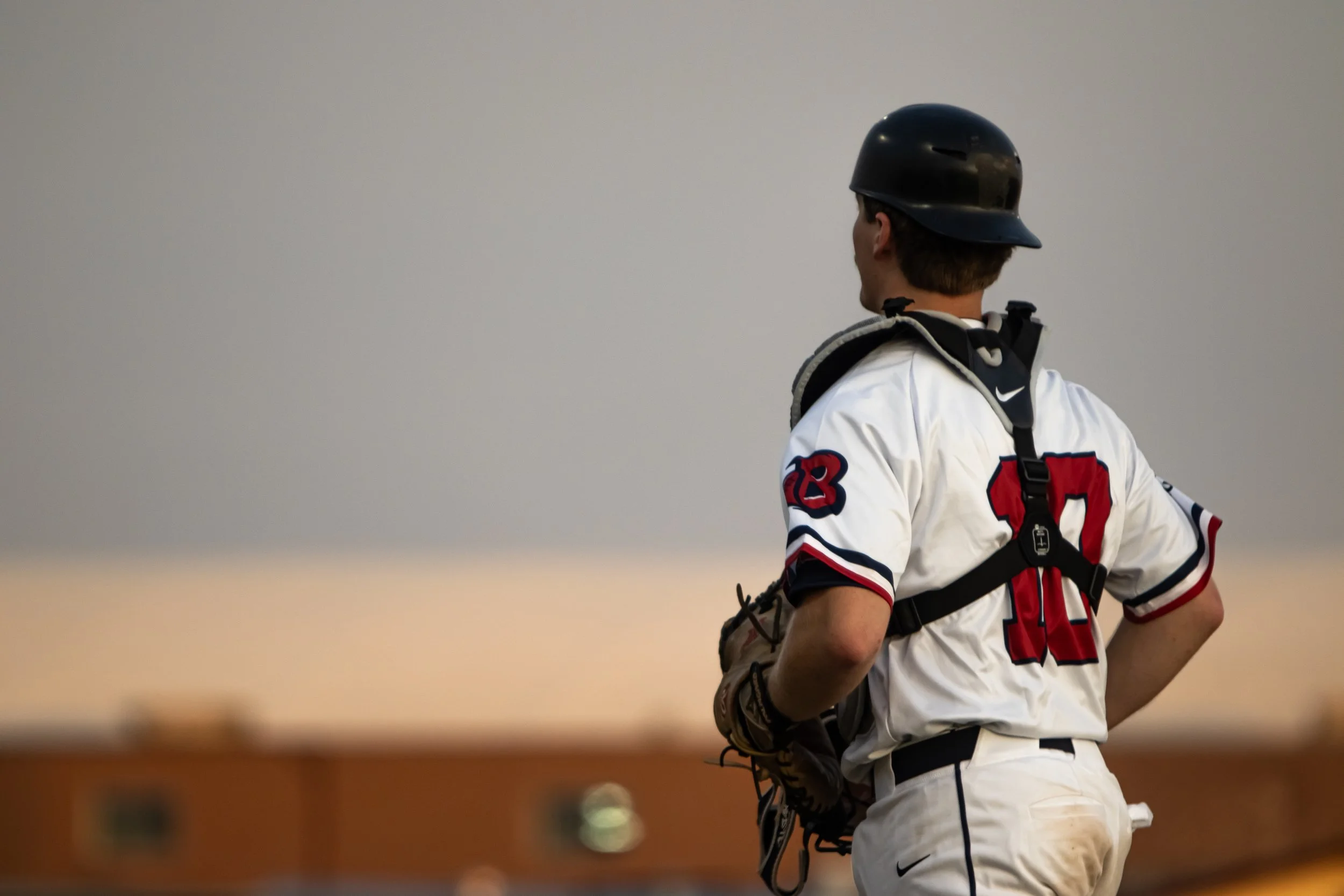 A baseball player wearing a black helmet and white jersey with red and black accents, standing on a field during sunset.