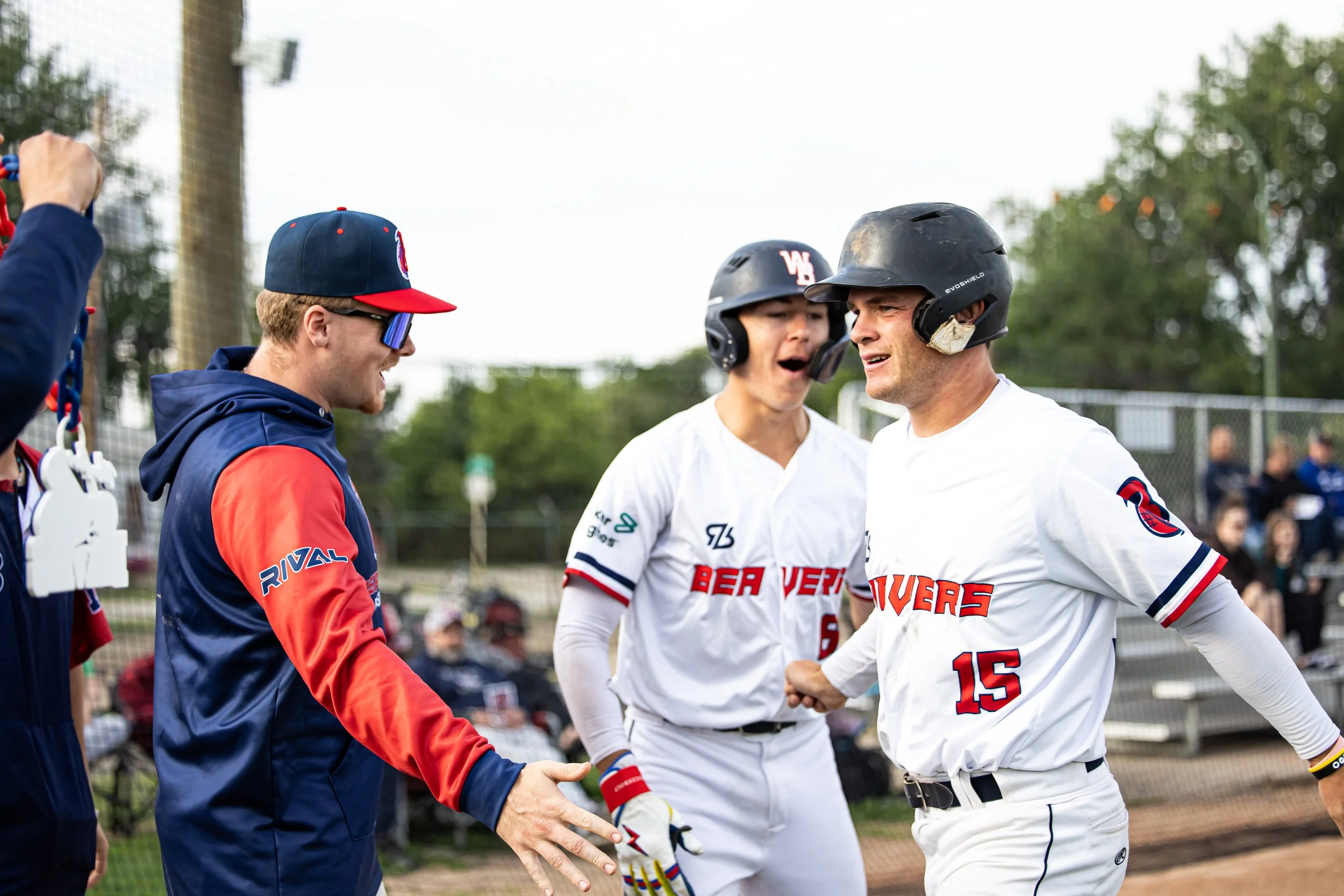 Three baseball players and a coach celebrating on the field. Two players wear black helmets and white uniforms, one of whom is shaking hands with a coach. Another player is dressed in a blue and red jacket and a black cap, smiling.