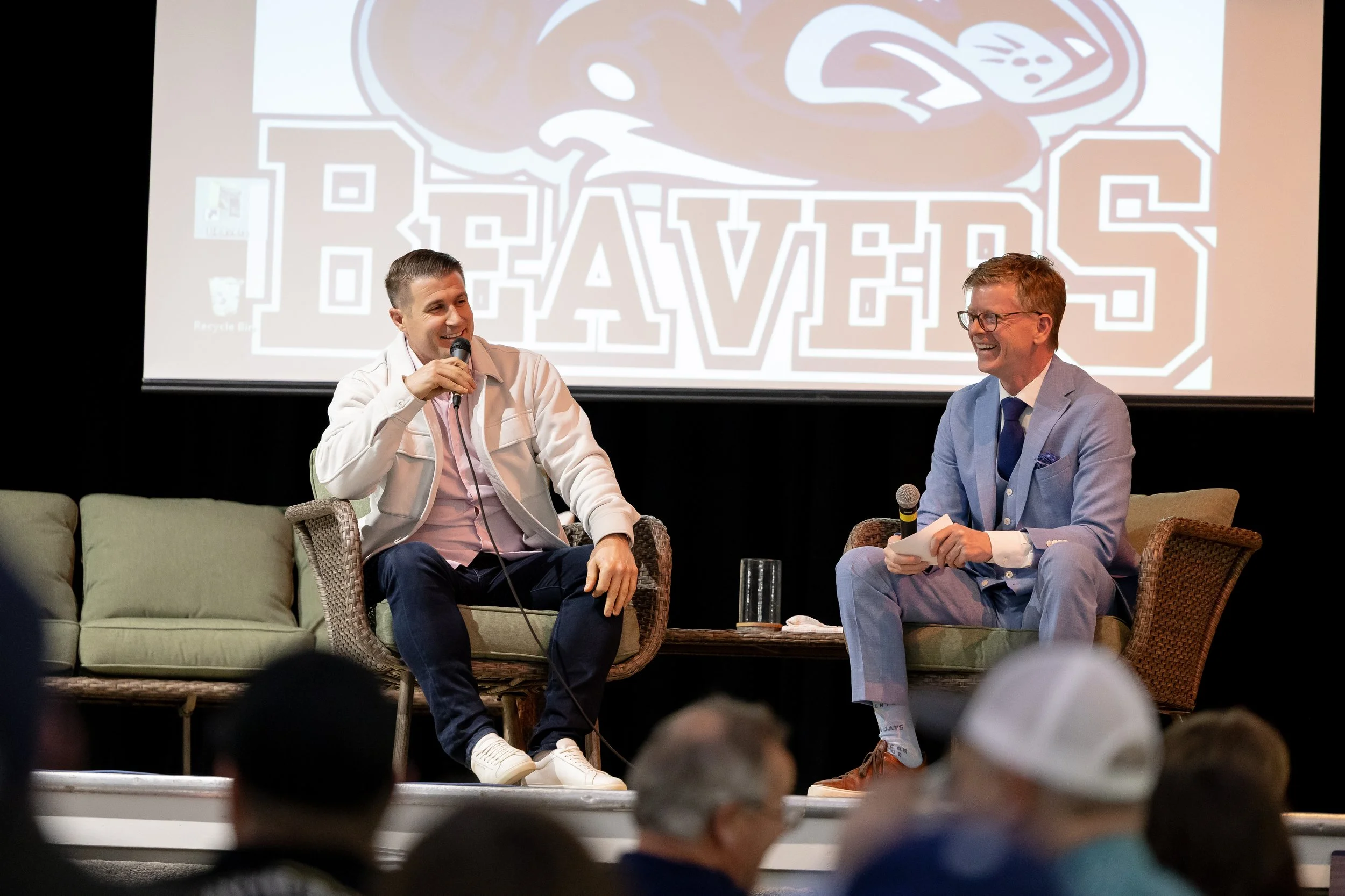 Two men are sitting on a stage, engaged in a conversation. They are holding microphones and smiling. Behind them is a large screen displaying the word 'BRAVE' with a graphic of a bird. Audience members are visible at the bottom of the image.