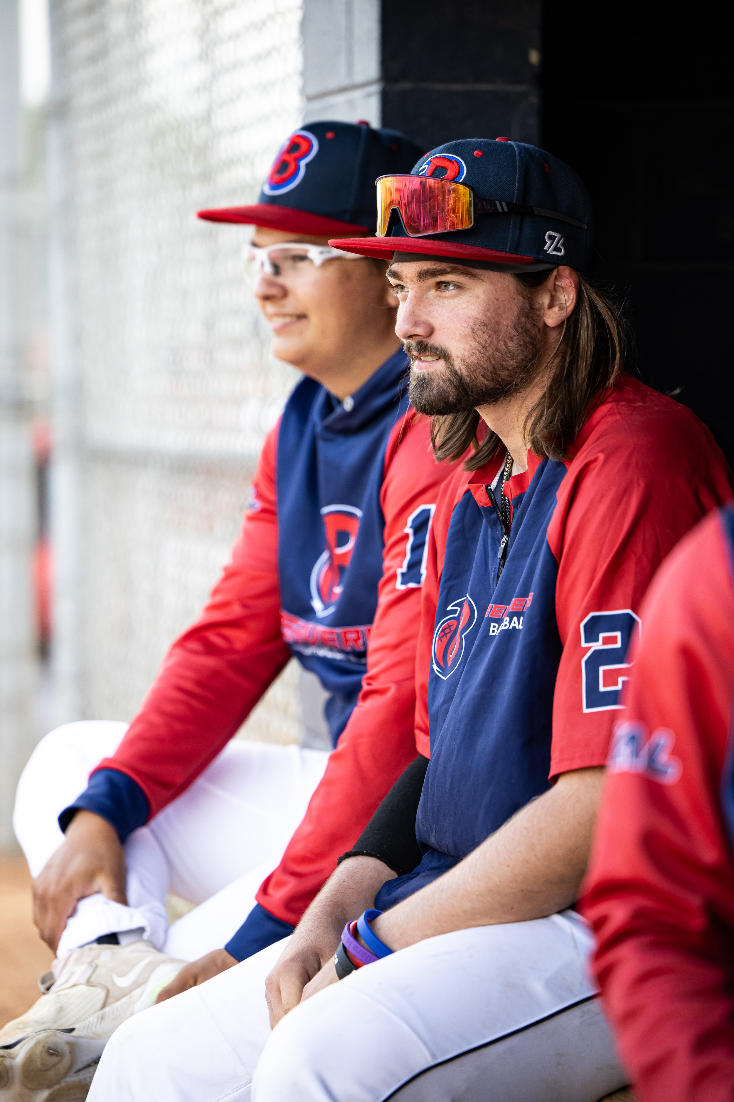Two baseball players sitting on a bench, wearing team jerseys and caps, with one player wearing sunglasses and the other wearing a sports watch.