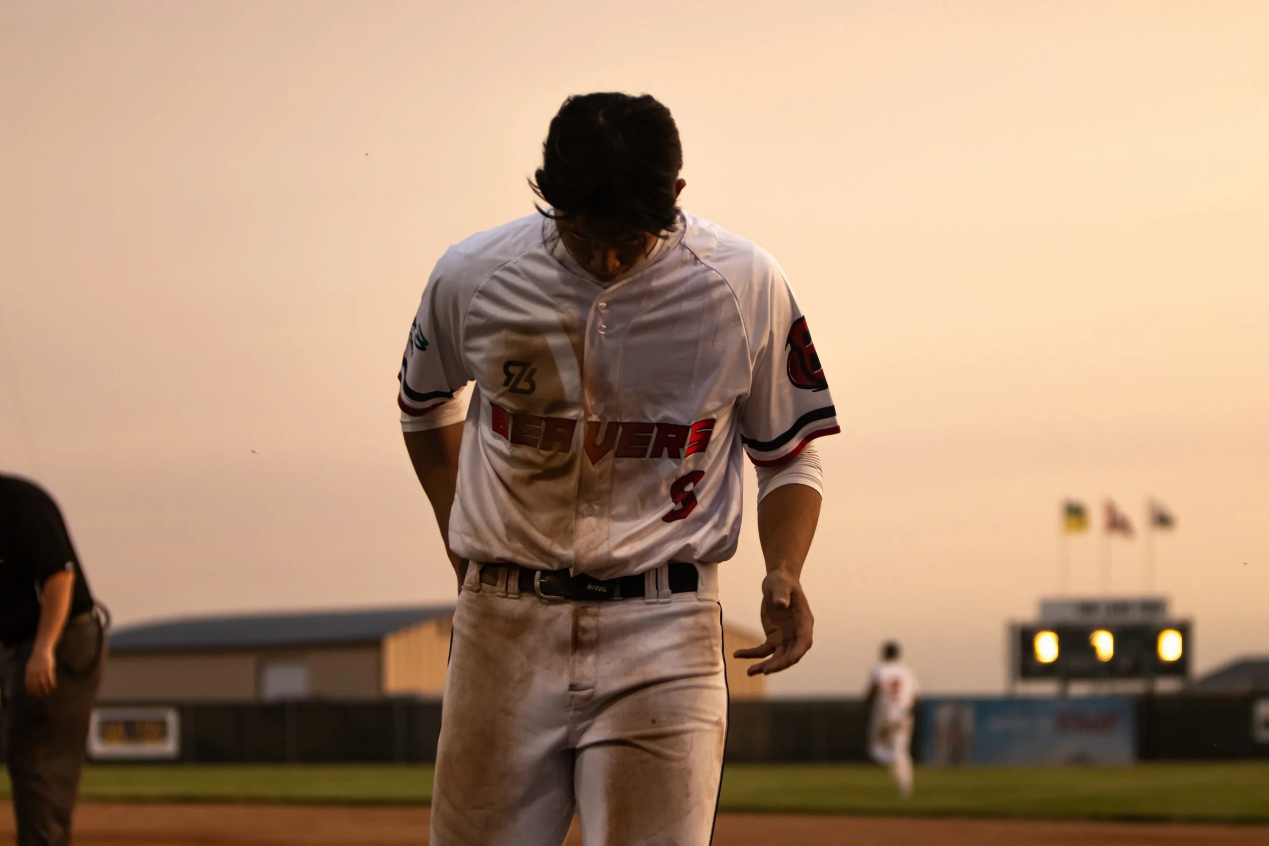 A baseball player dressed in a white jersey and khaki pants, looking down with a serious expression in a stadium during what appears to be evening or night. Other players can be seen in the background.