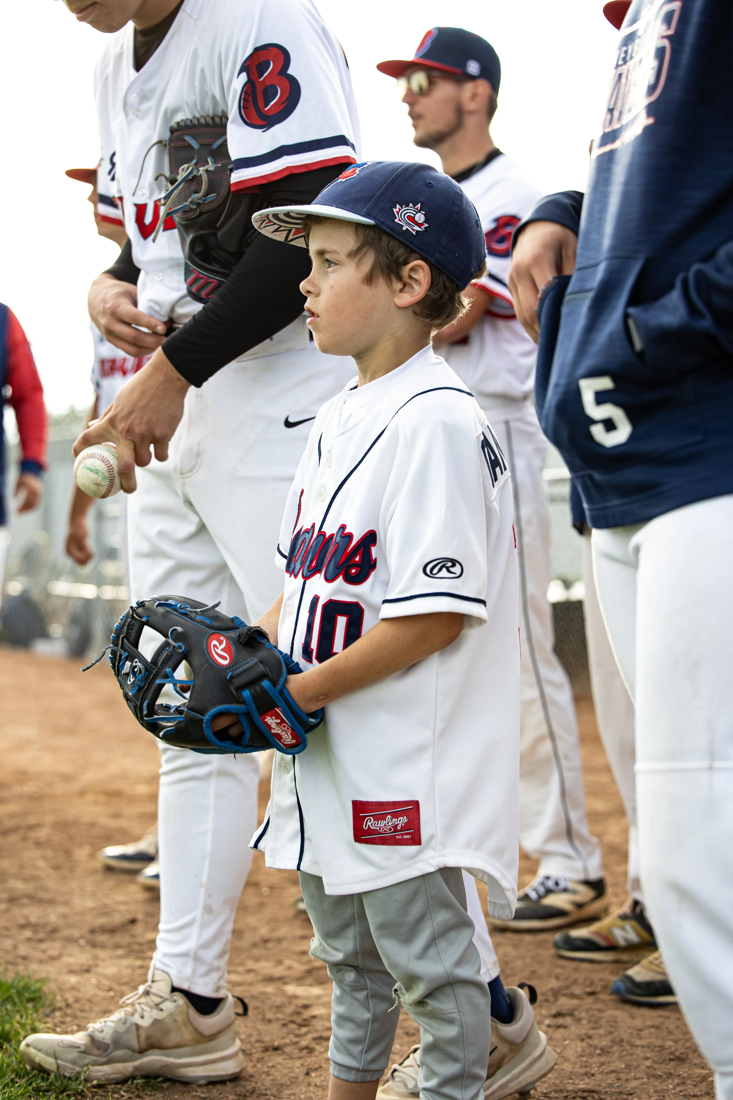 A young boy wearing a baseball cap, baseball jersey, and glove standing on a baseball field during a team lineup with adults and other players.