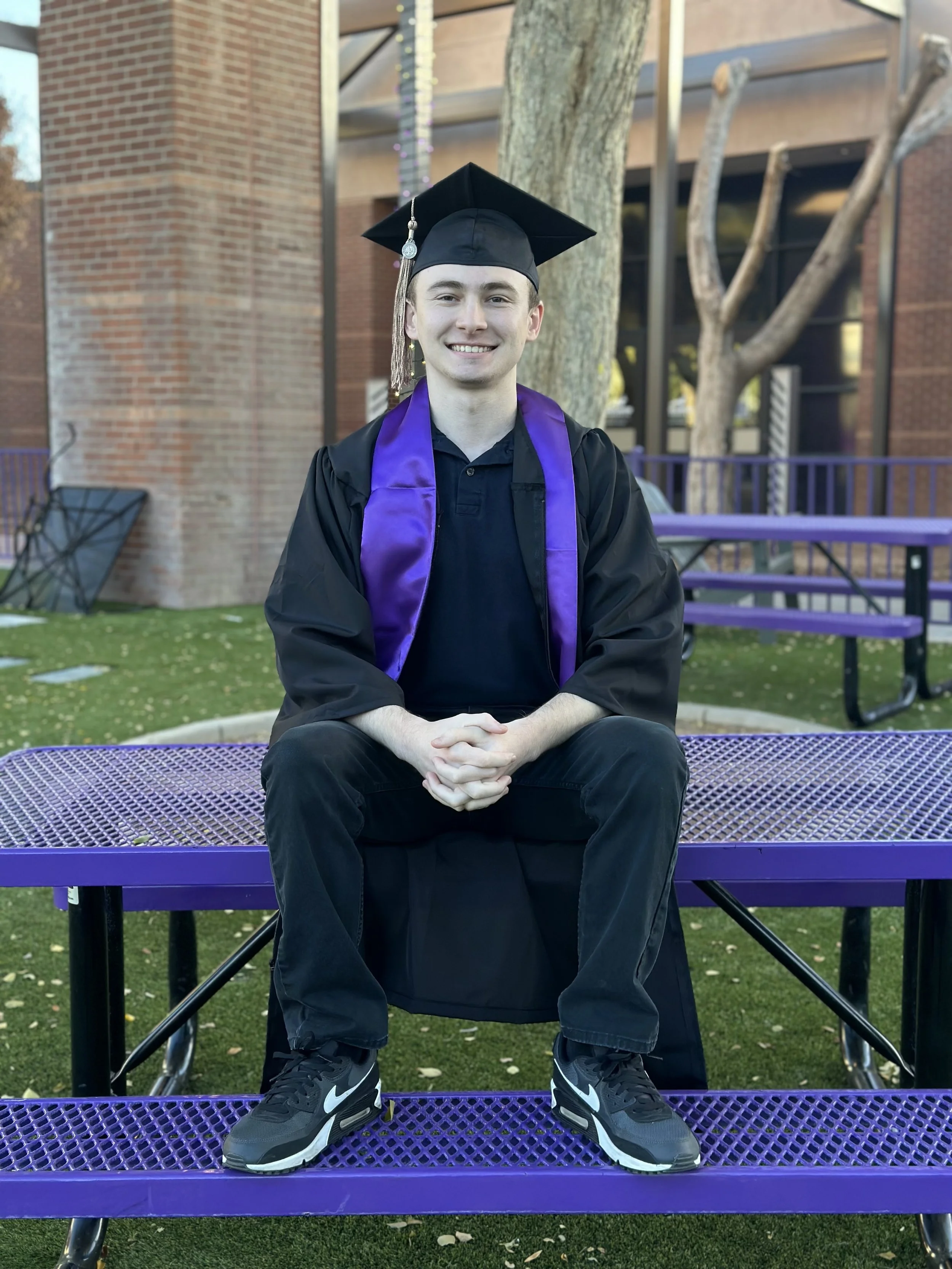 A young man in graduation attire sitting on a purple bench outdoors. He is wearing a black cap and gown with a purple sash, and black sneakers with white Nike logos. There are trees and a brick building in the background.