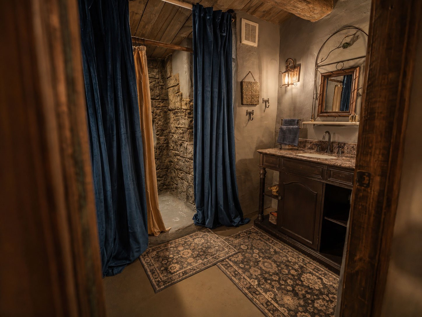 Rustic hobbit bathroom at Mirador Texas with a stone shower, dark wood vanity with marble countertop, framed mirror, vintage wall sconce, and patterned rugs.