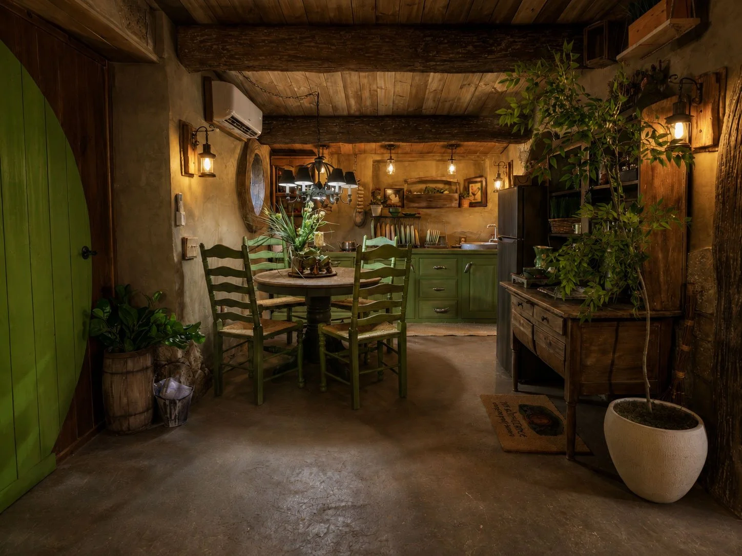 Cozy rustic hobbit-inspired kitchen with wooden ceiling beams, green cabinets, a round dining table with four green chairs, potted plants, vintage furniture, and warm lighting.