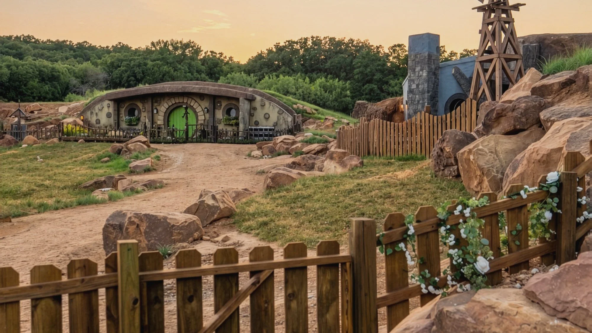 Sunset over Mirador's Texas Shire near Mingus, Texas: a dirt lane leads to the Gardener House's round green door, with the Writer House's chimney and windmill beside red sandstone and a fence wrapped in white flowers