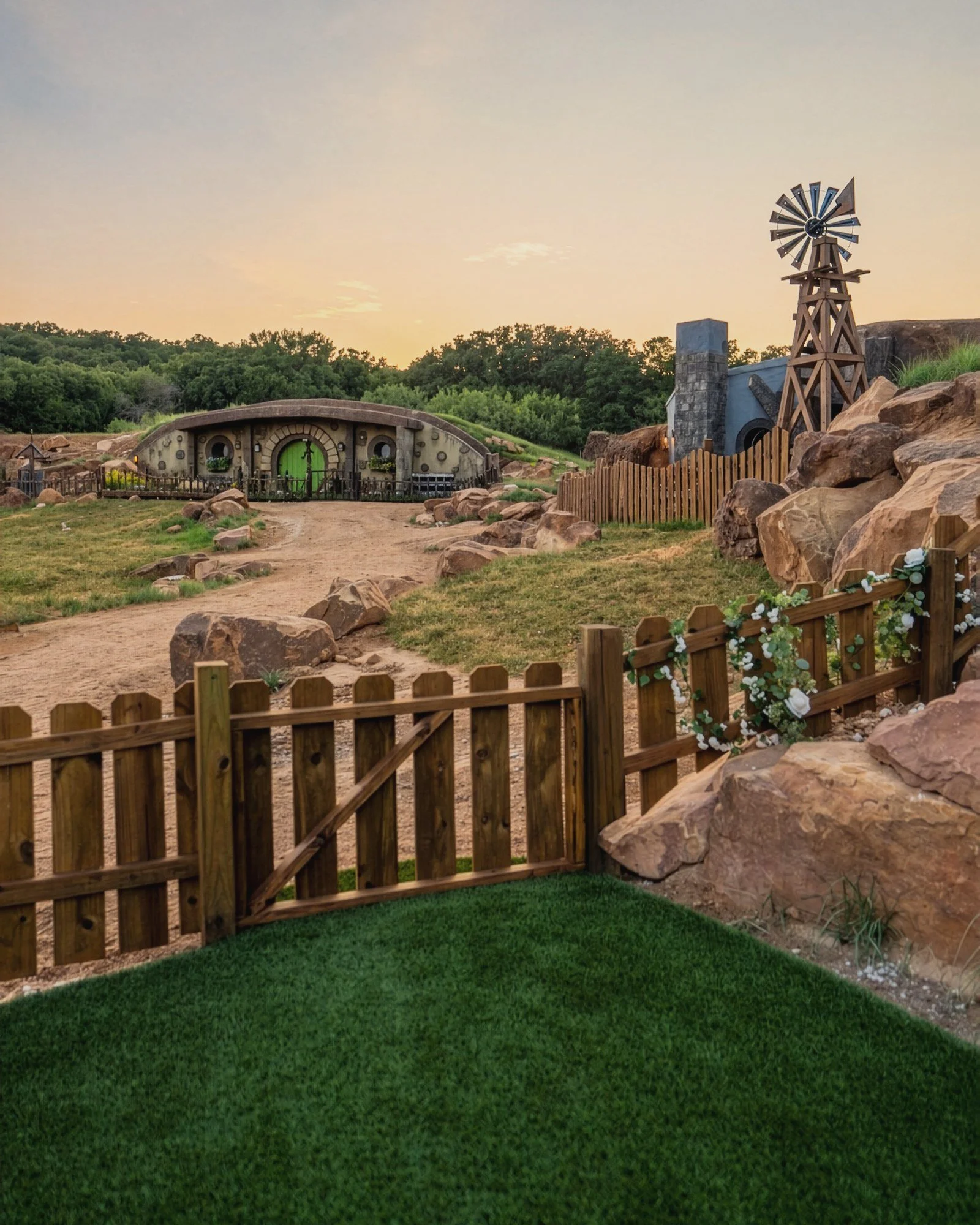 A wooden gate with a white flower garland at sunset, looking down a dirt lane to the Gardener House's round green door, with the Writer House's chimney and windmill at right
