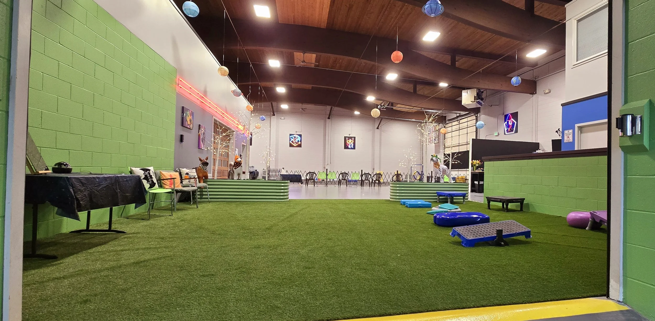 Indoor play area with green artificial grass, decorated with colorful paper lanterns hanging from the wooden ceiling, and cartoon animal statues, including a giraffe and two zebras. There are chairs, small tables, and various small toys and play equipment scattered around.