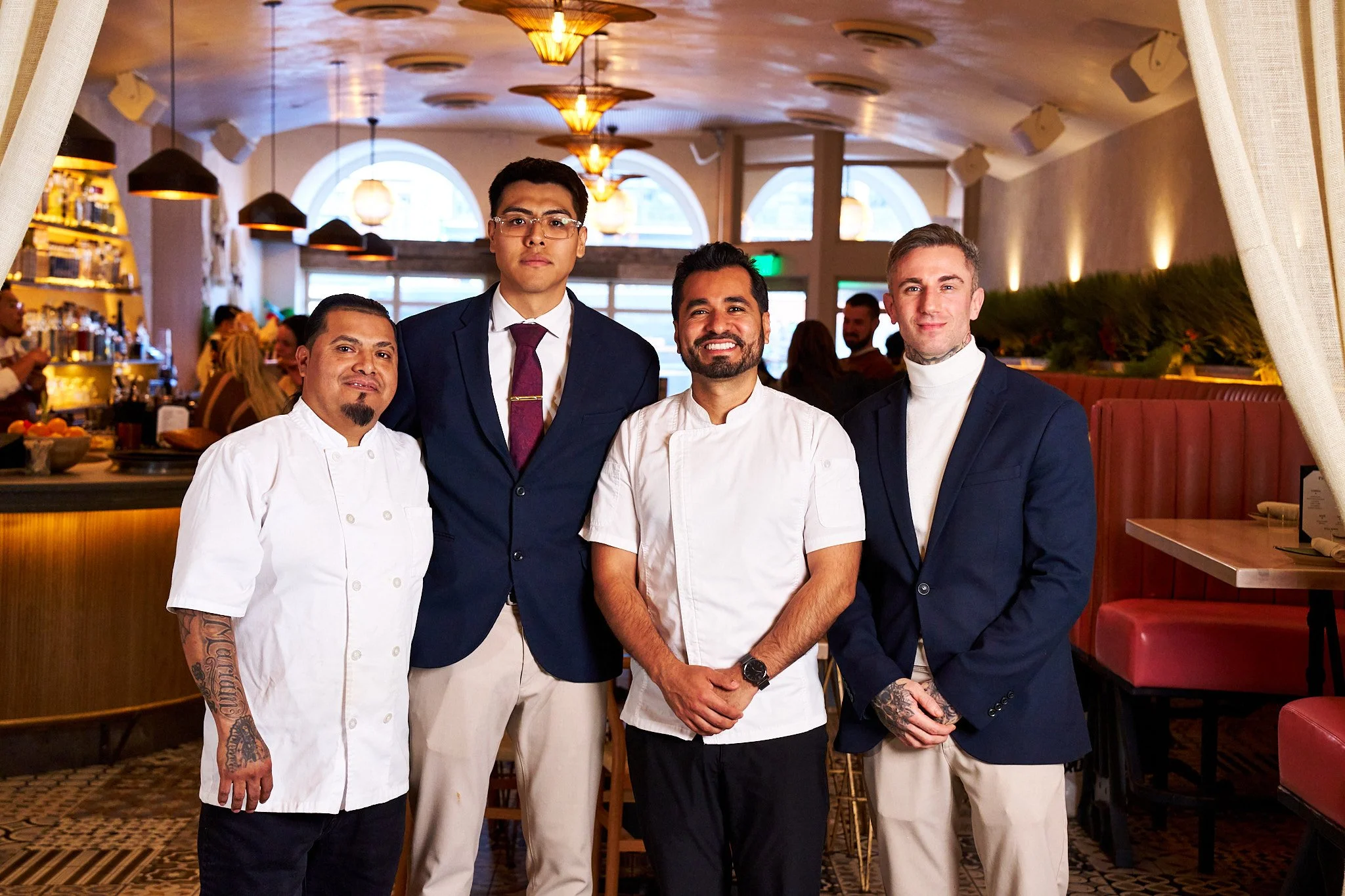 Four men standing together inside a restaurant. Two are dressed as chefs in white uniforms, and two are dressed in suits. The background shows restaurant decor with warm lighting, arched windows, and a bar area.