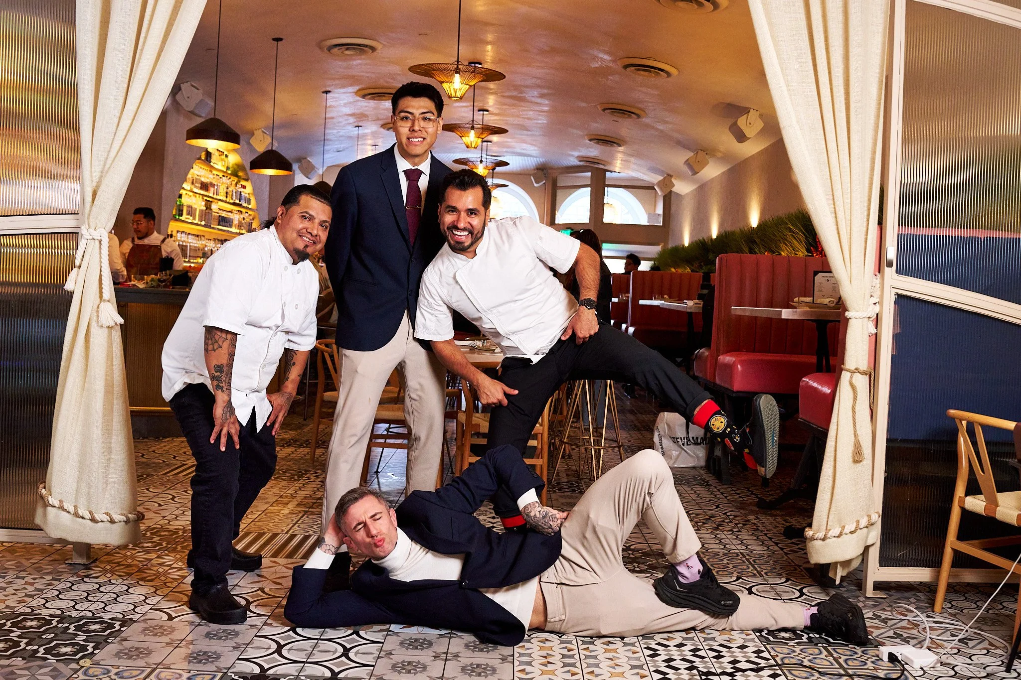 Group of six men posing for a photo inside a restaurant. They are dressed in a mix of casual and formal attire, with some in chef uniforms and one in a suit. They are all smiling or making playful expressions, with some standing and some sitting or lying on the floor. The restaurant has warm lighting, patterned floor tiles, and a shelf with bottles in the background.
