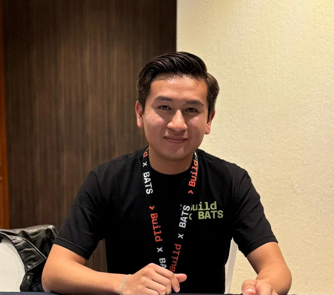Joven hombre con cabello oscuro peinado hacia atrás, sonriente, usando camiseta negra con letras verdes y blancas, y collar con letras blancas y rojas, sentado en una mesa en un cuarto con pared beige y pared de madera en el fondo.