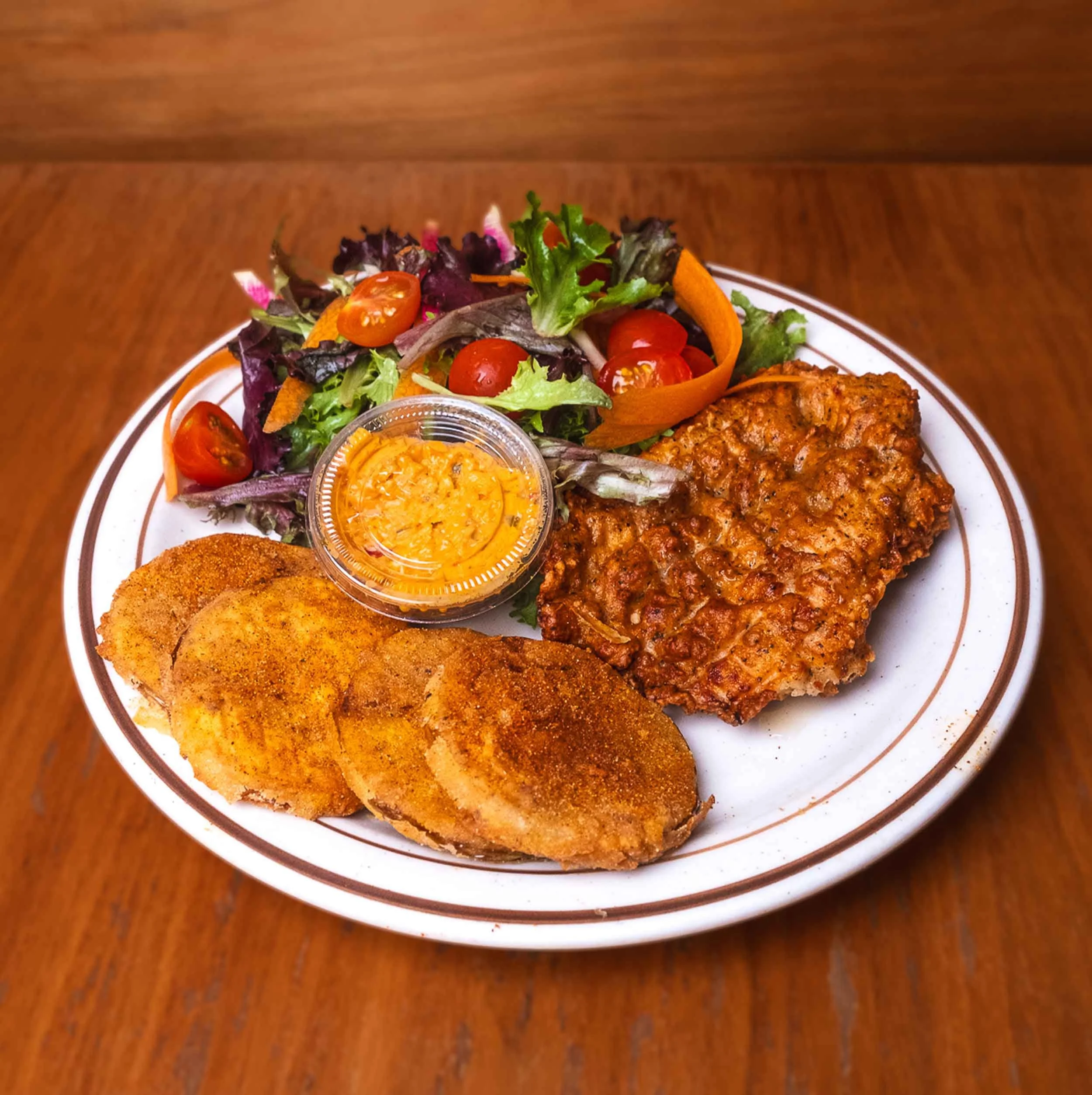 A plate with fried fish fillets, a side salad with cherry tomatoes, lettuce, and shredded vegetables, and a small container of creamy orange dipping sauce, all on a wooden table.