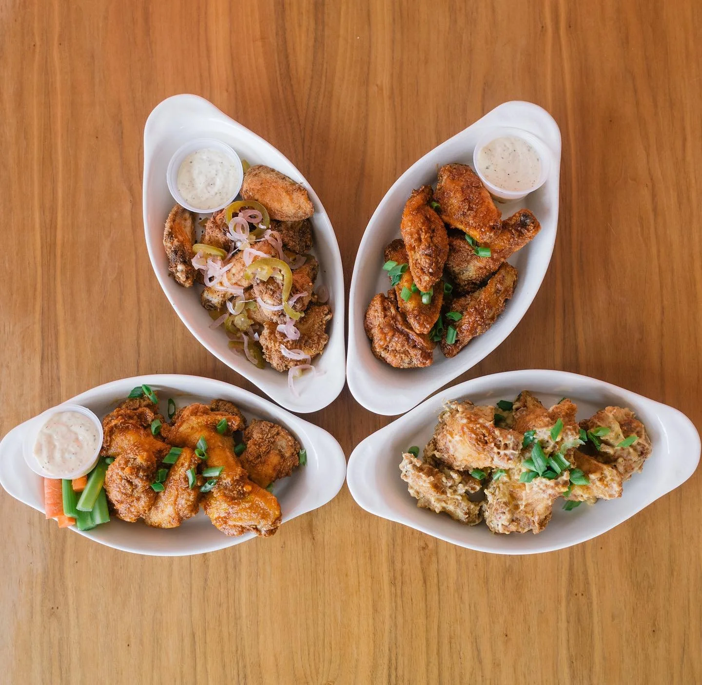 Four white oval dishes of fried chicken with different sauces and garnishes on a wooden table.