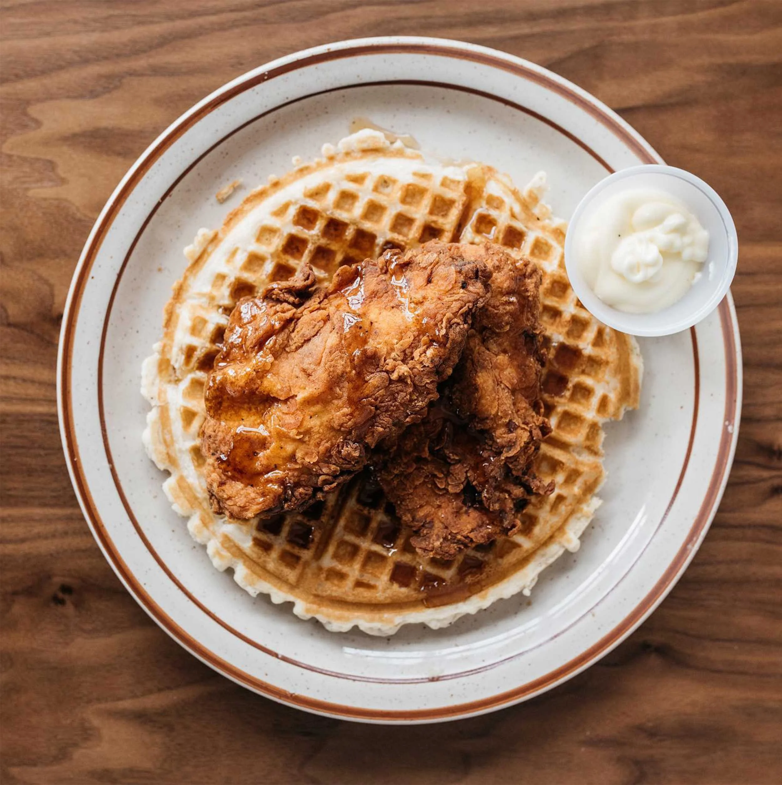 Plate with two waffles, fried chicken, and a cup of white gravy, on a wooden table.