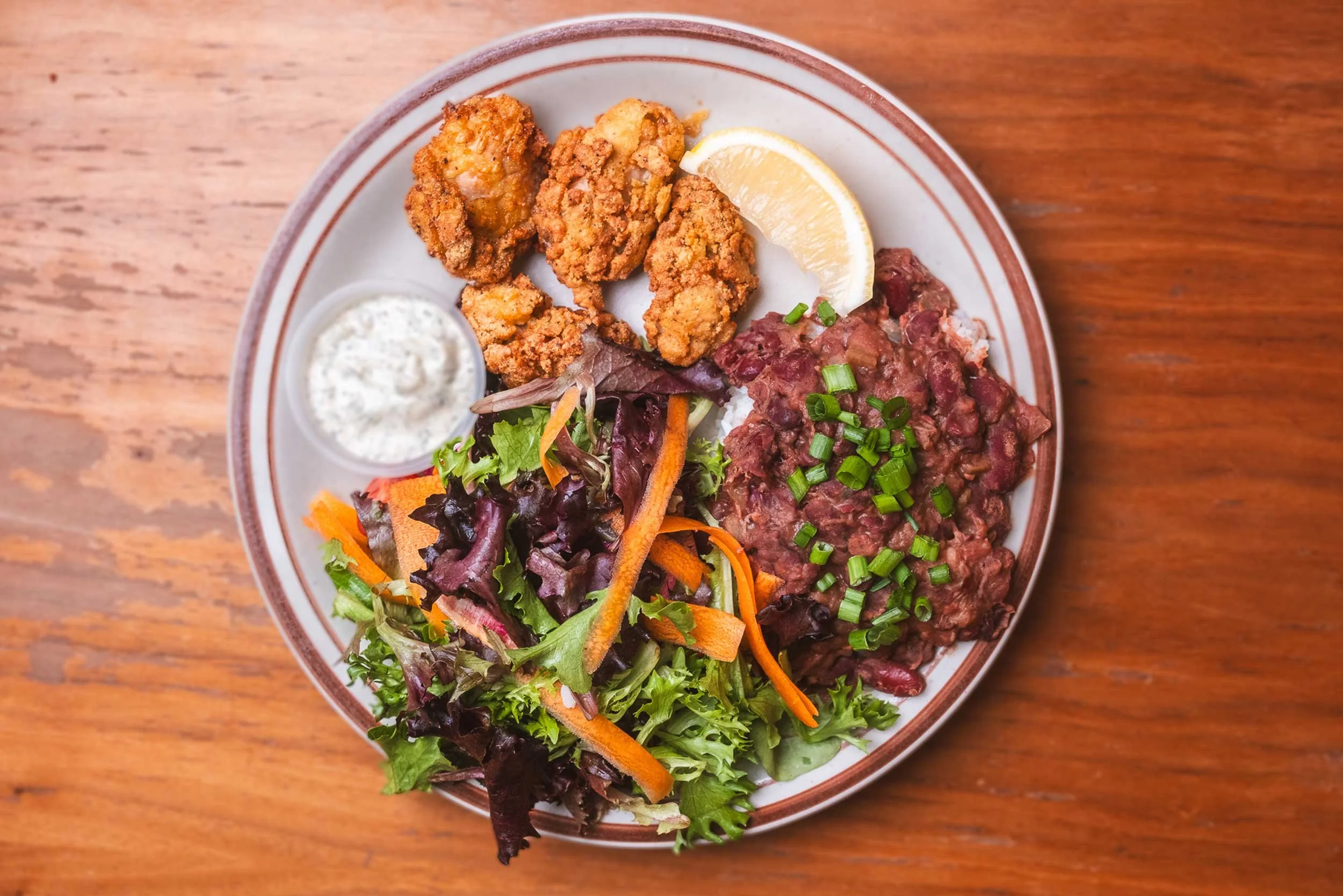 A plate of fried chicken tenders, a lemon wedge, a small cup of creamy dipping sauce, a serving of baked beans topped with chopped green onions, and a colorful side salad with mixed greens, shredded carrots, and purple lettuce, on a wooden table.