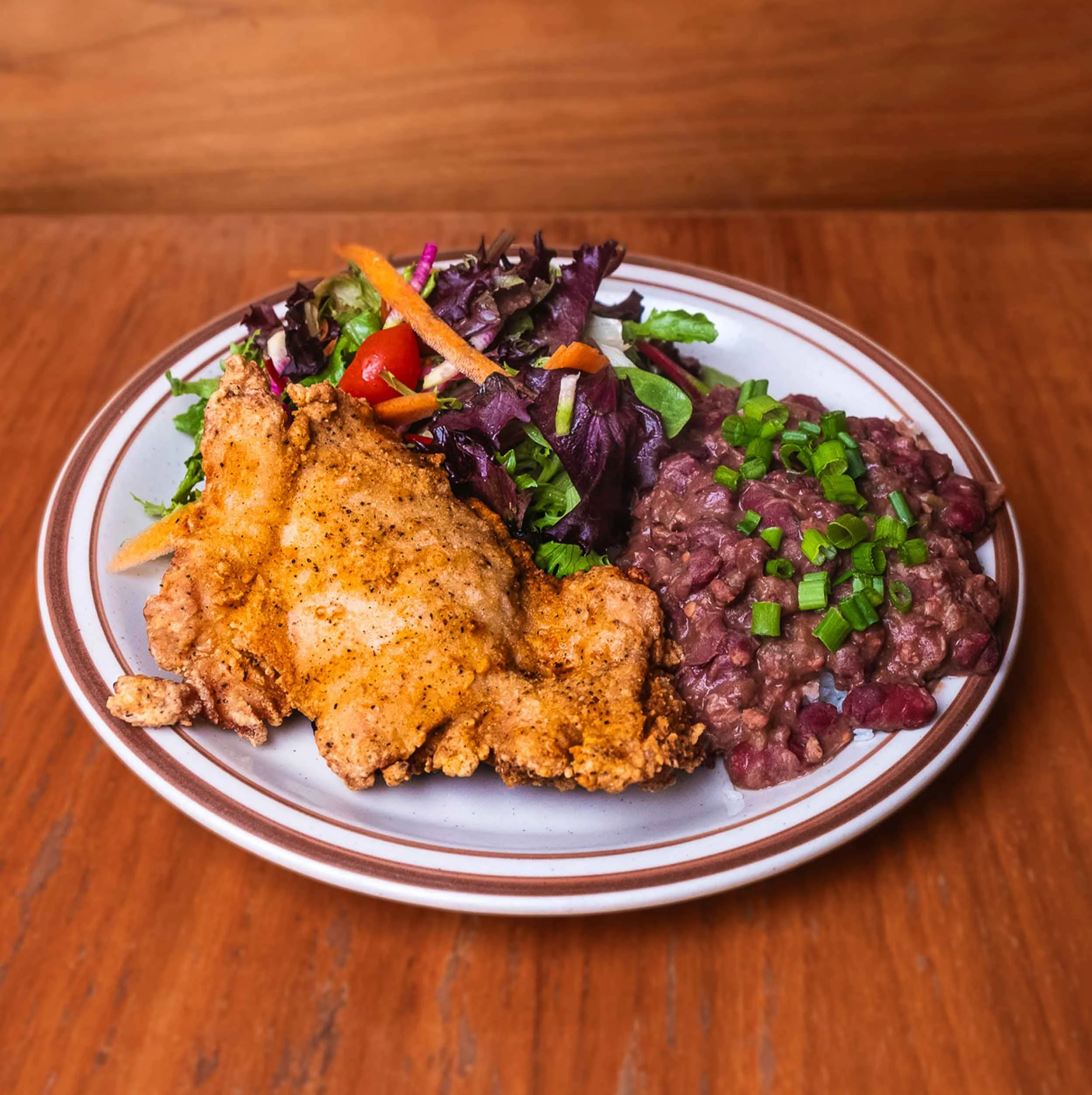 Close-up of a plate with a grilled chicken fillet, mixed greens, cherry tomatoes, shredded carrots, and black beans on a wooden table.