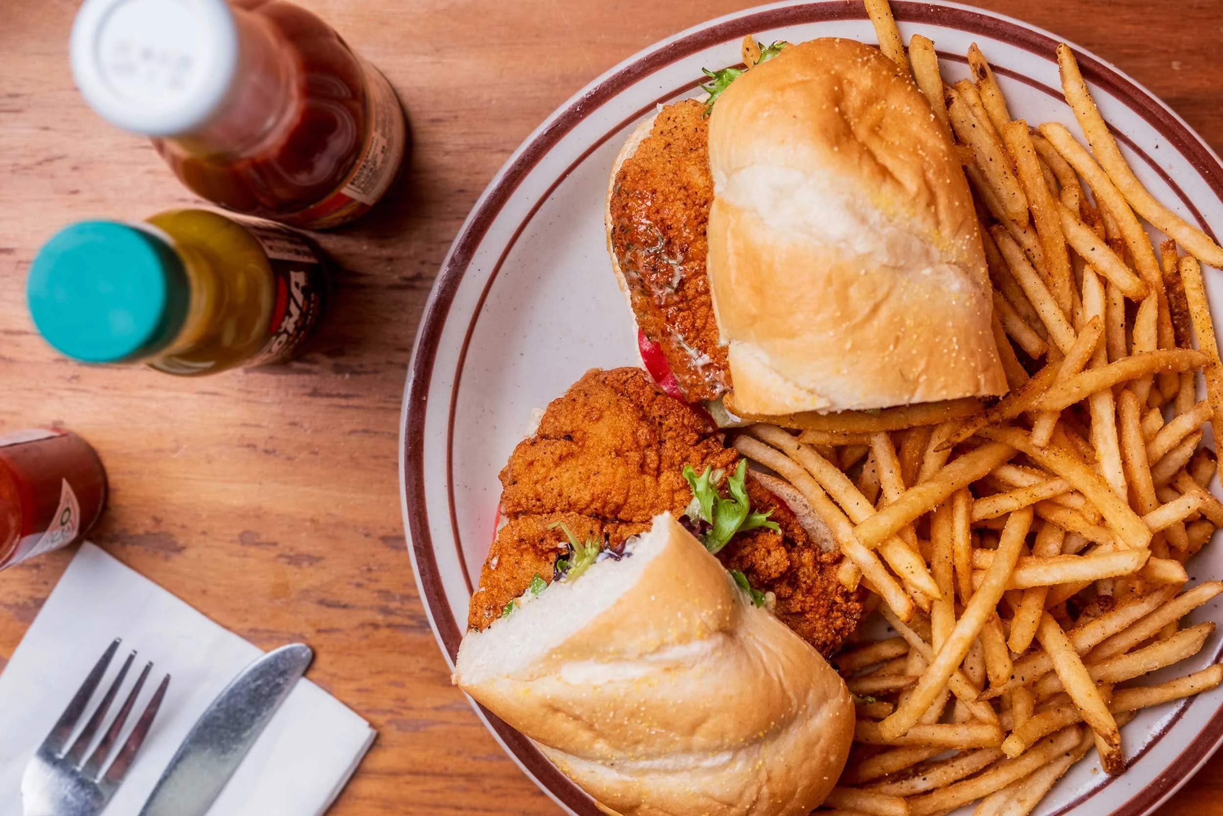 Plate of French fries, fried chicken, and a sandwich bun on a wooden table with condiments and utensils nearby.
