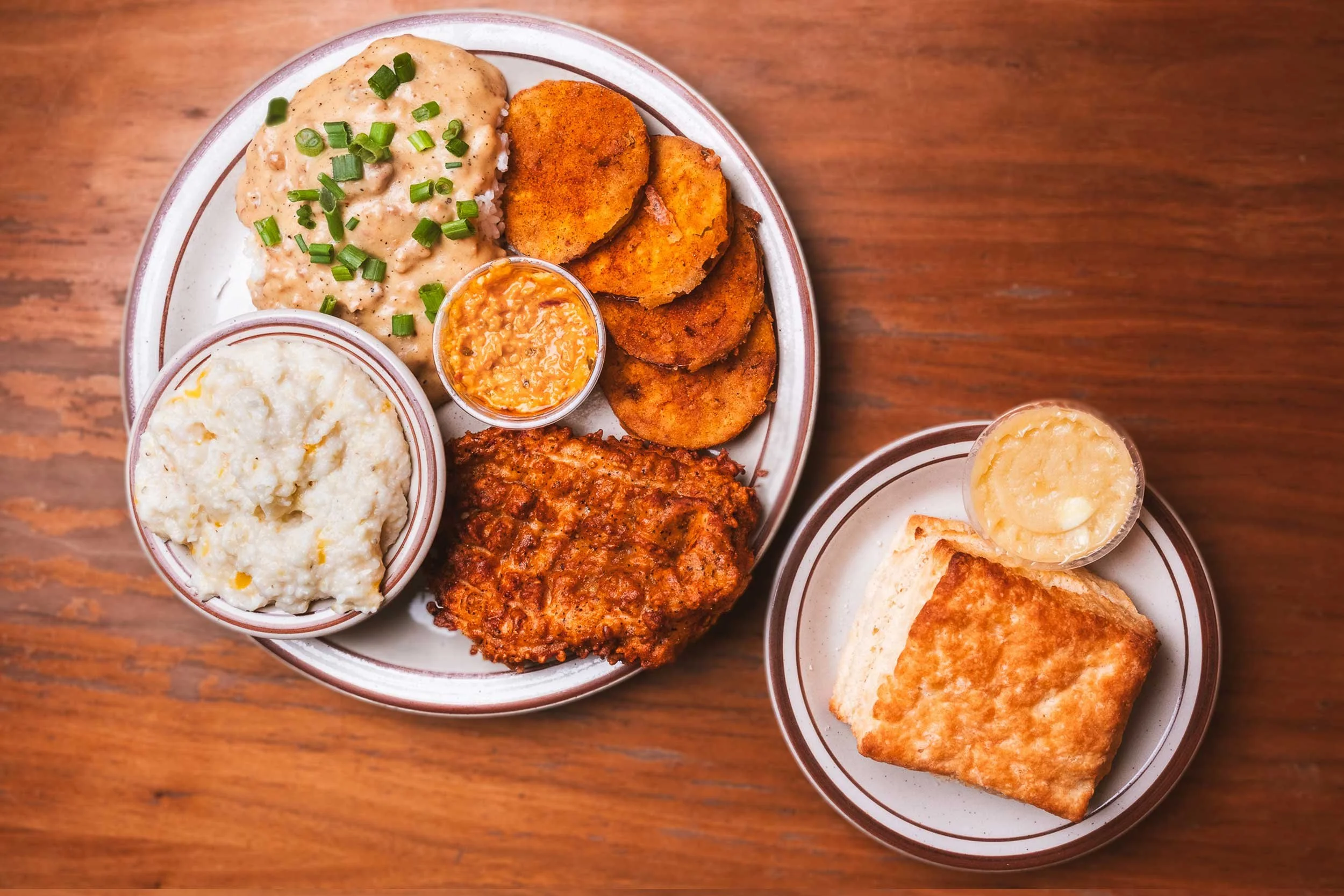 Plate of Southern comfort food including mashed potatoes, chicken fried steak, fried green tomatoes, macaroni and cheese, and a bowl of sauce, served on a wooden table.