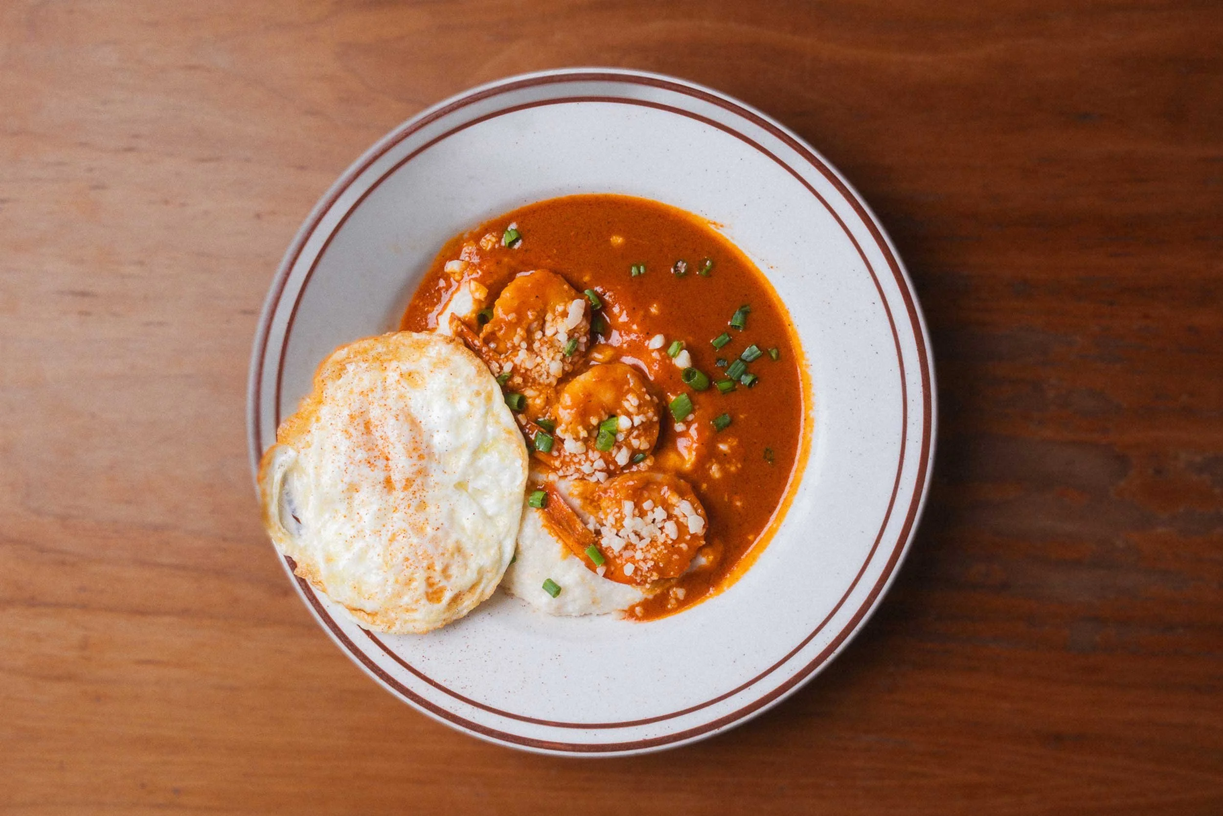 A bowl of shrimp and grits, topped with parsley, and served with a fried egg on the side, on a wooden table.