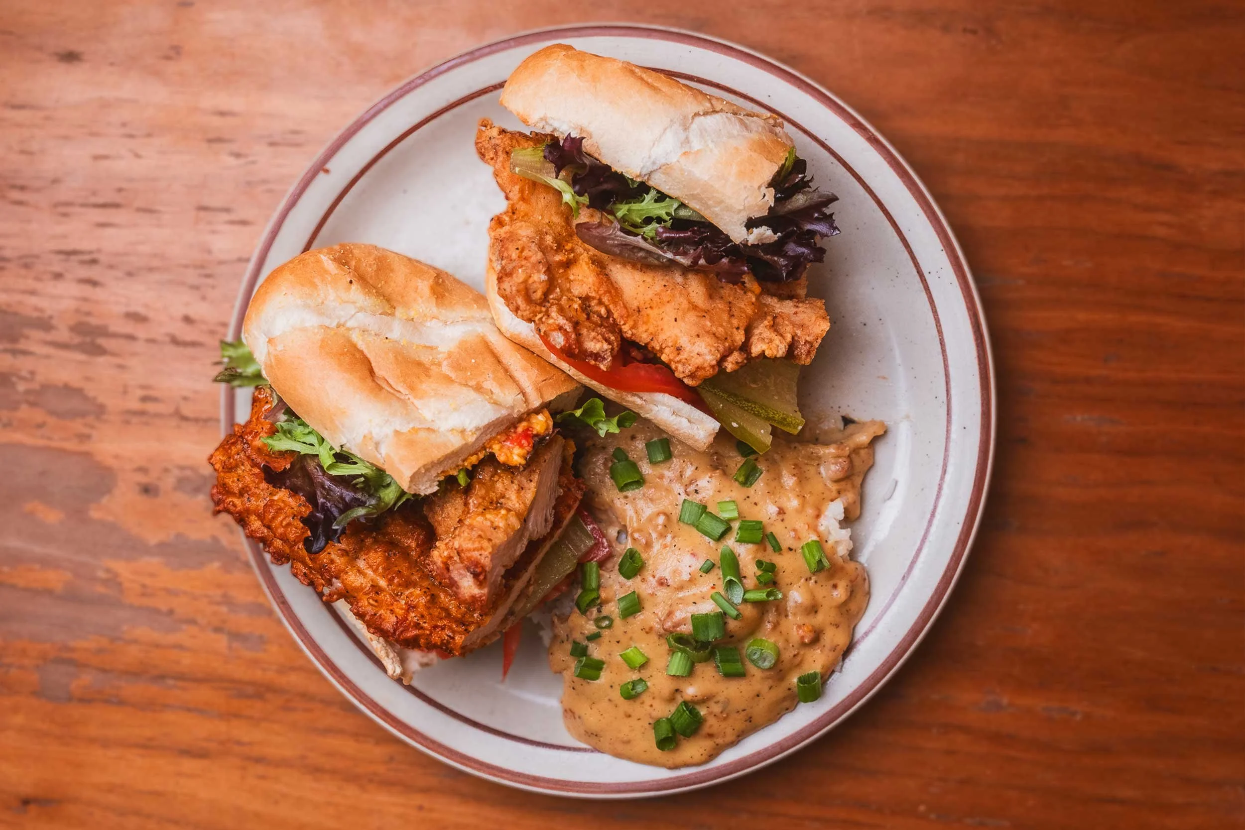 Plate holding two chicken sandwiches with lettuce and pickles, and a serving of creamy mashed potatoes with chopped green onions, on a wooden table.